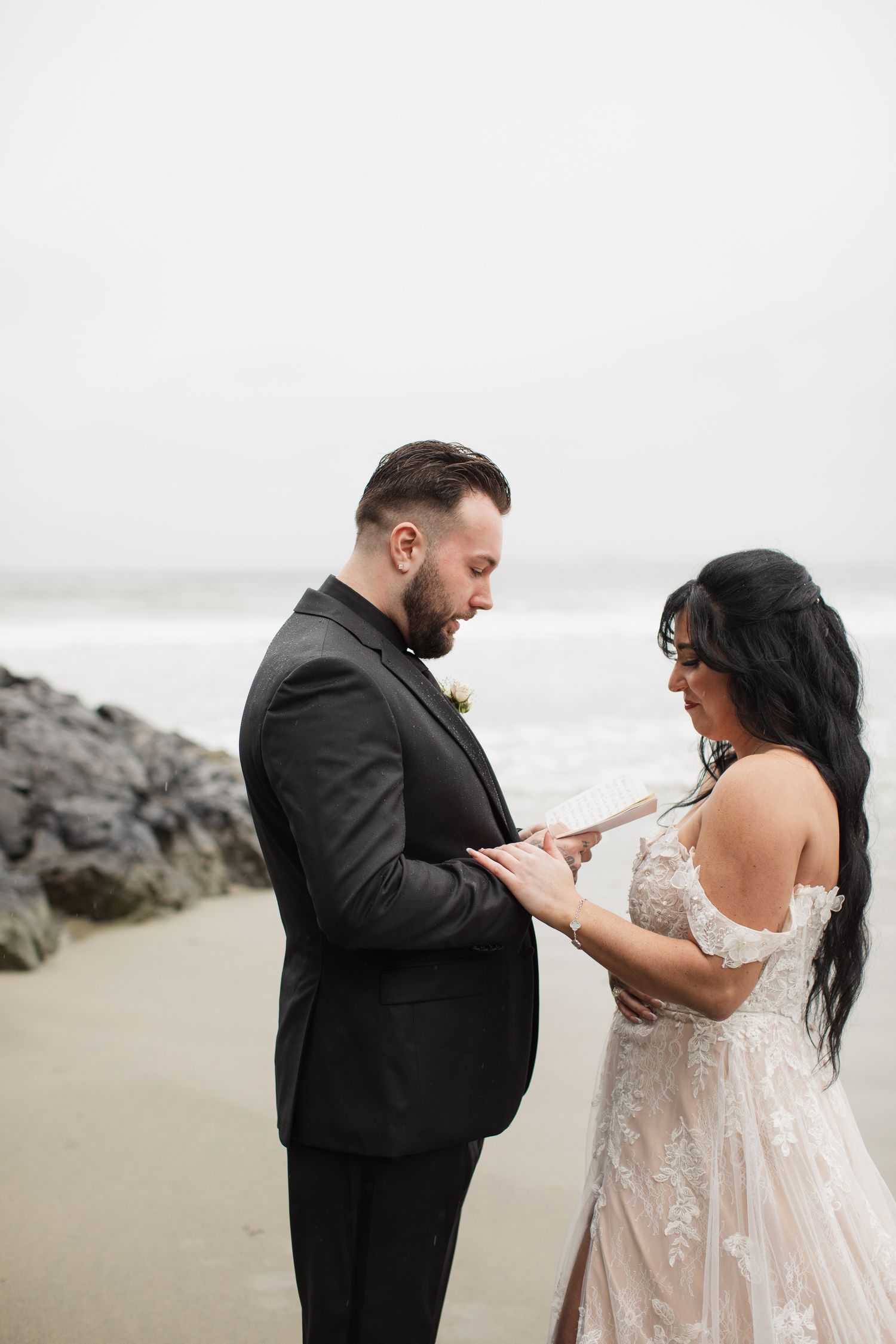 Bride and groom exchange vows on a misty beach, groom reading from paper while holding hands.