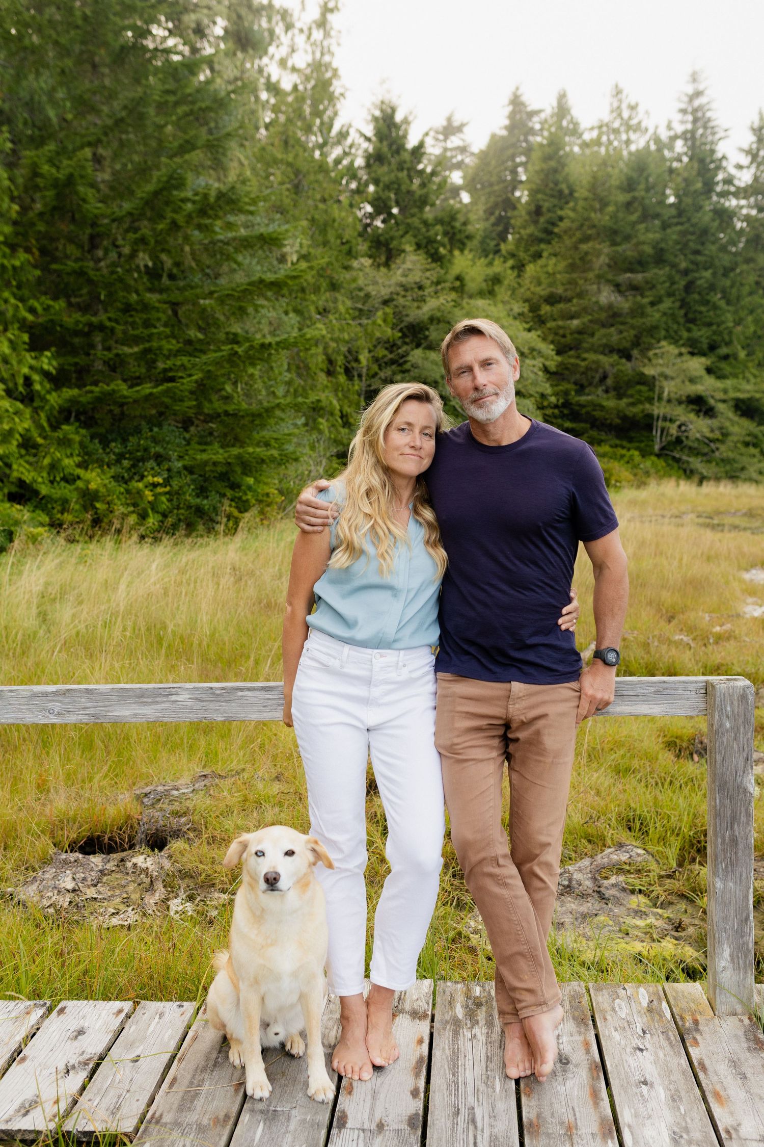Happy couple posing with their golden retriever on a weathered wooden dock in a scenic meadow.