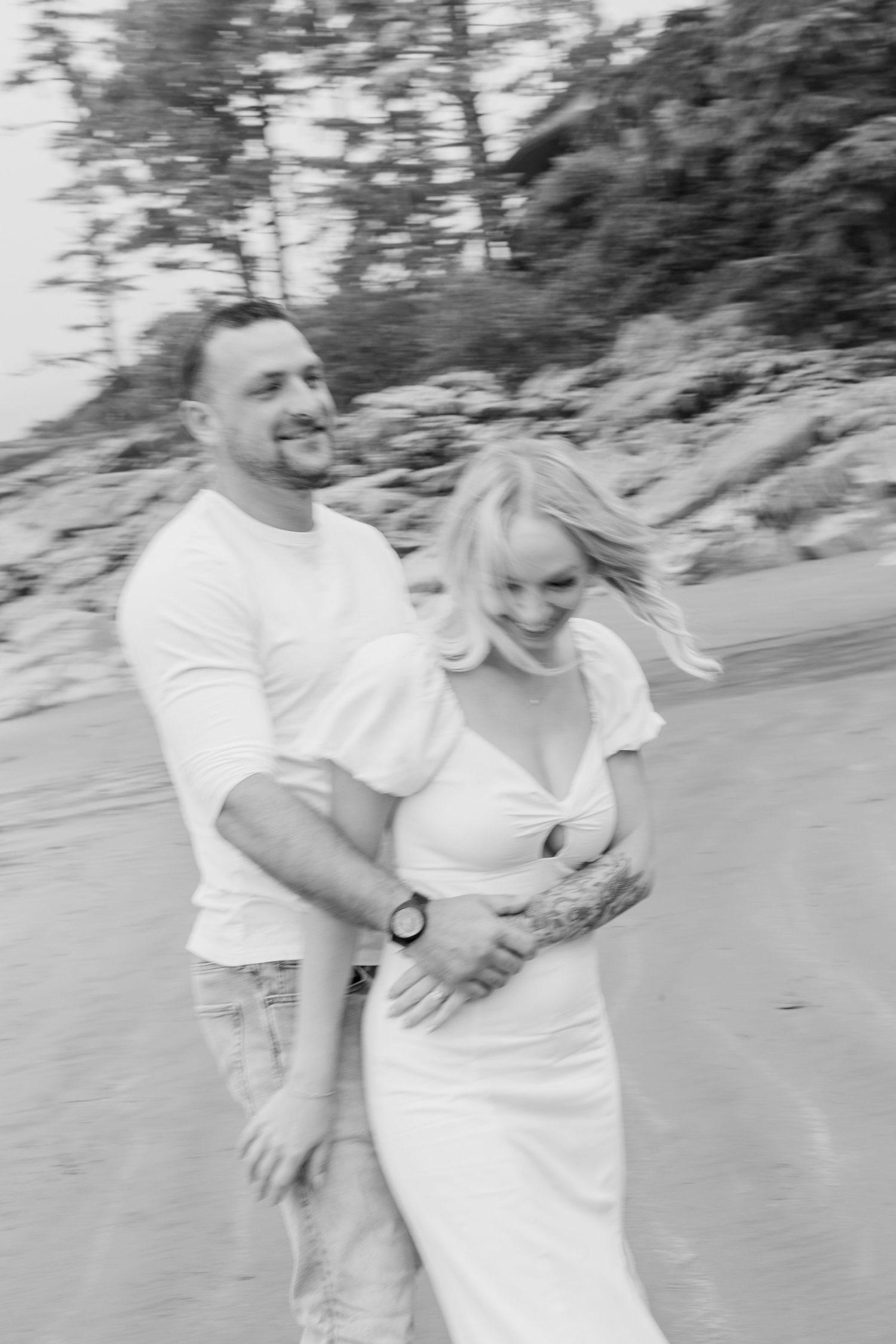 Black and white photo of laughing couple embracing while walking on windy beach.