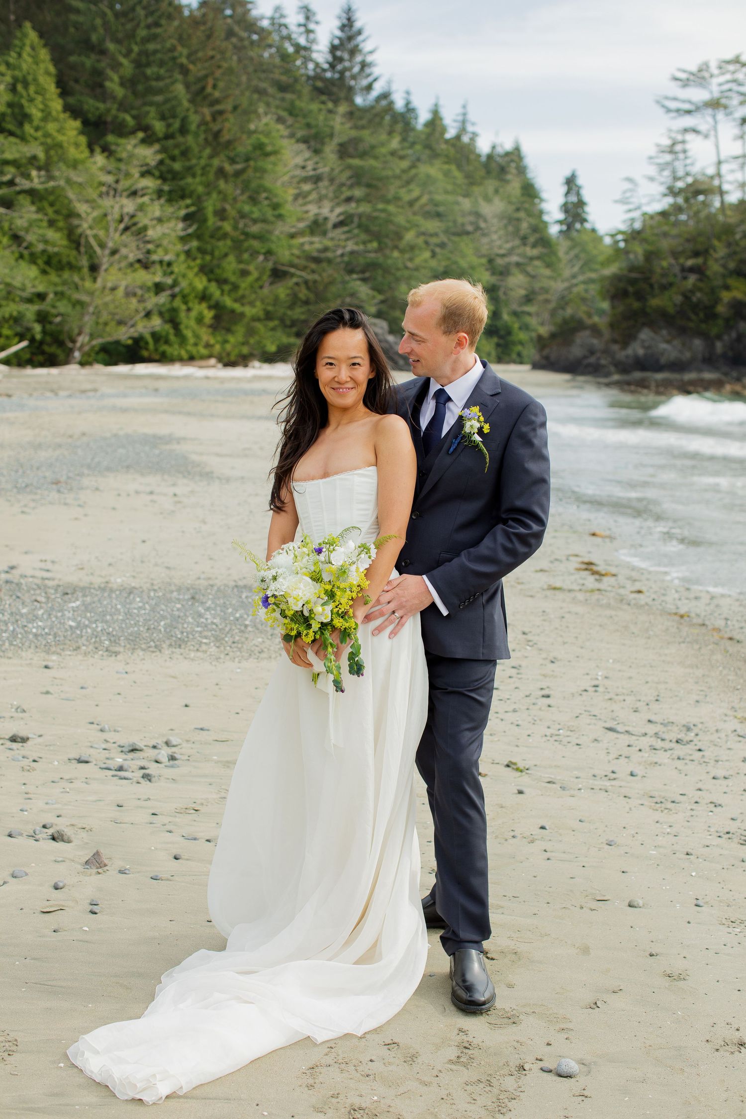Bride in white gown and groom in navy suit walk on sandy beach with forest backdrop.