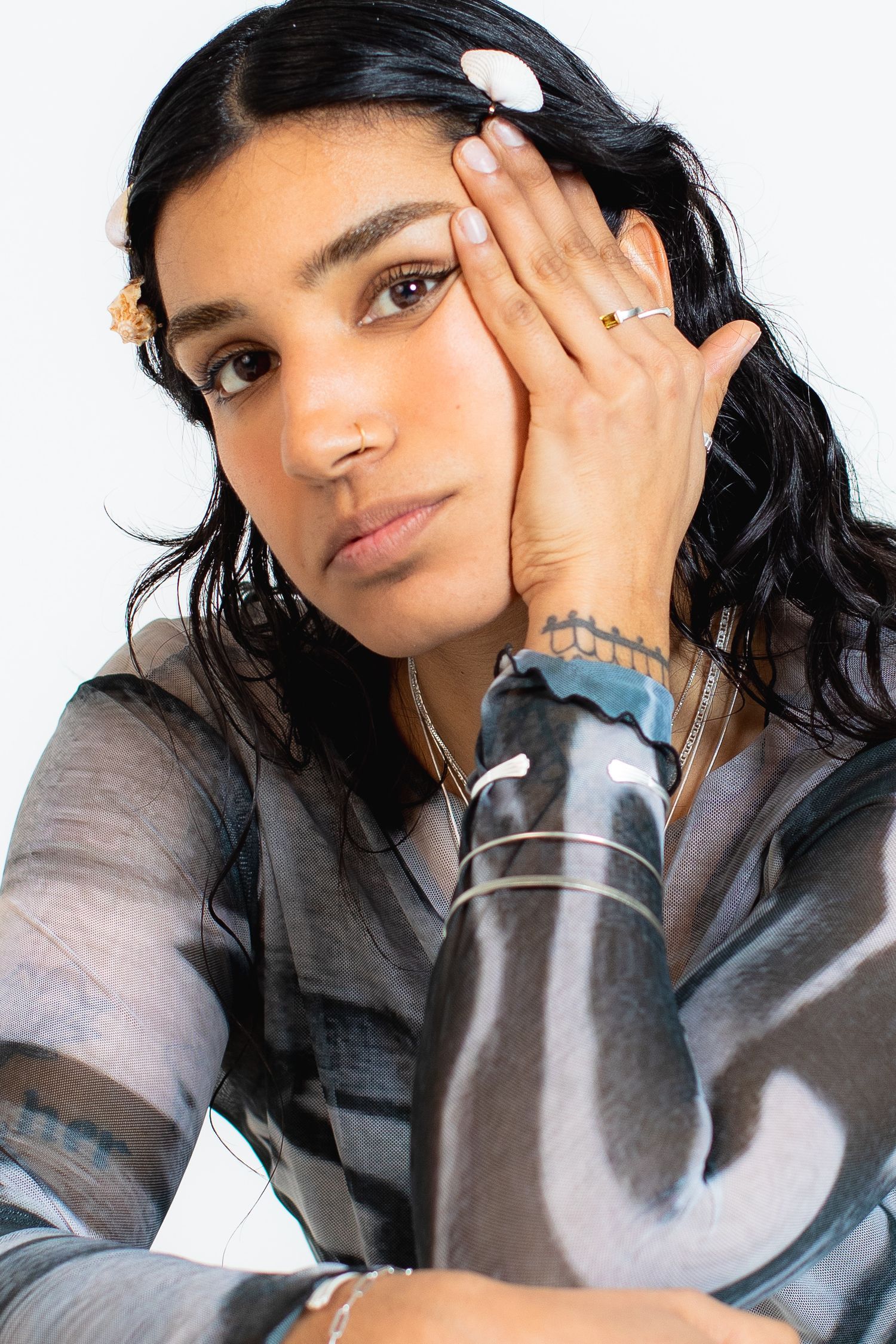 Young woman with dark wavy hair wearing sheer top and gold jewelry resting hand on face.