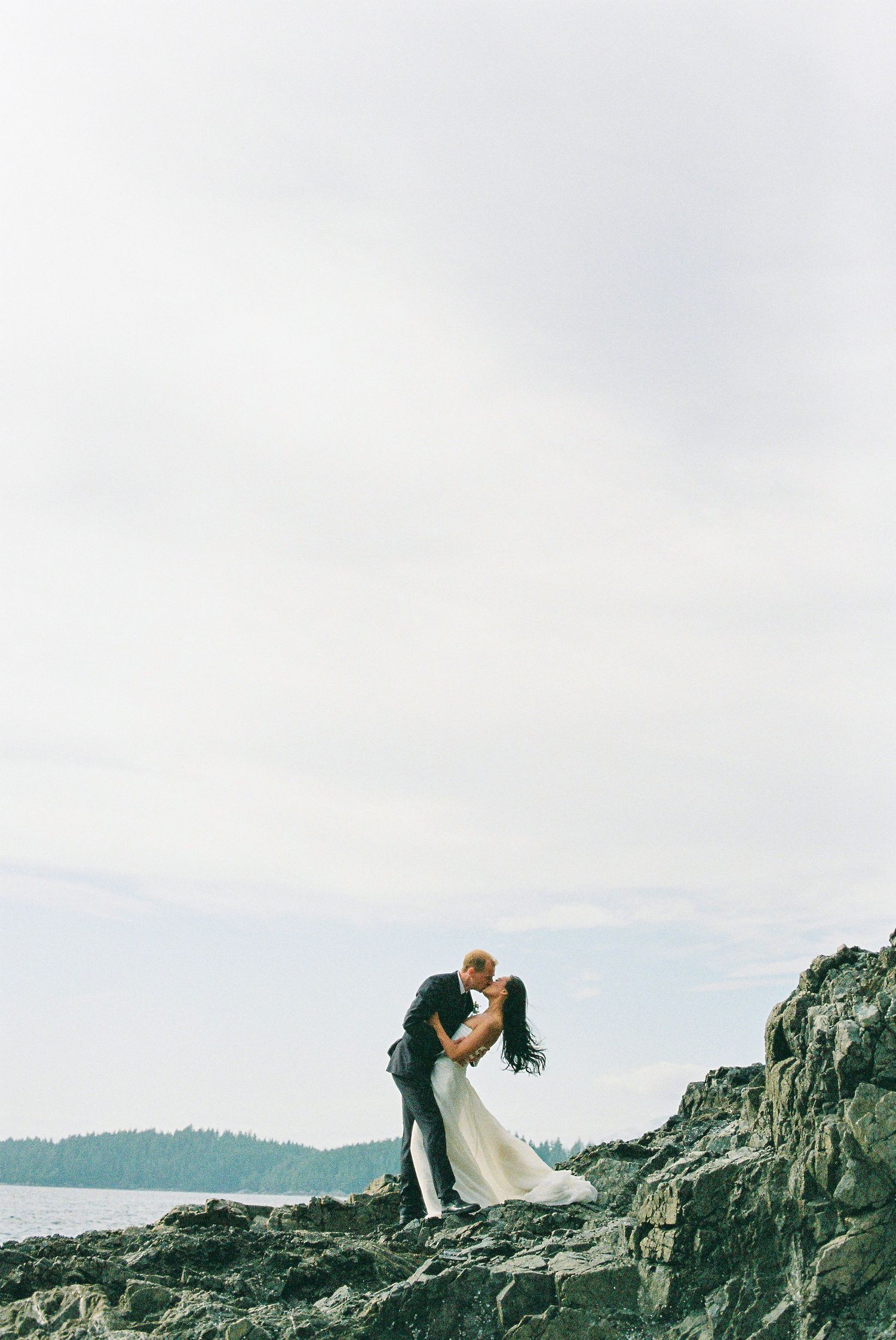 Bride and groom sharing a romantic kiss on rocky coastal shoreline with ocean and forest backdrop.