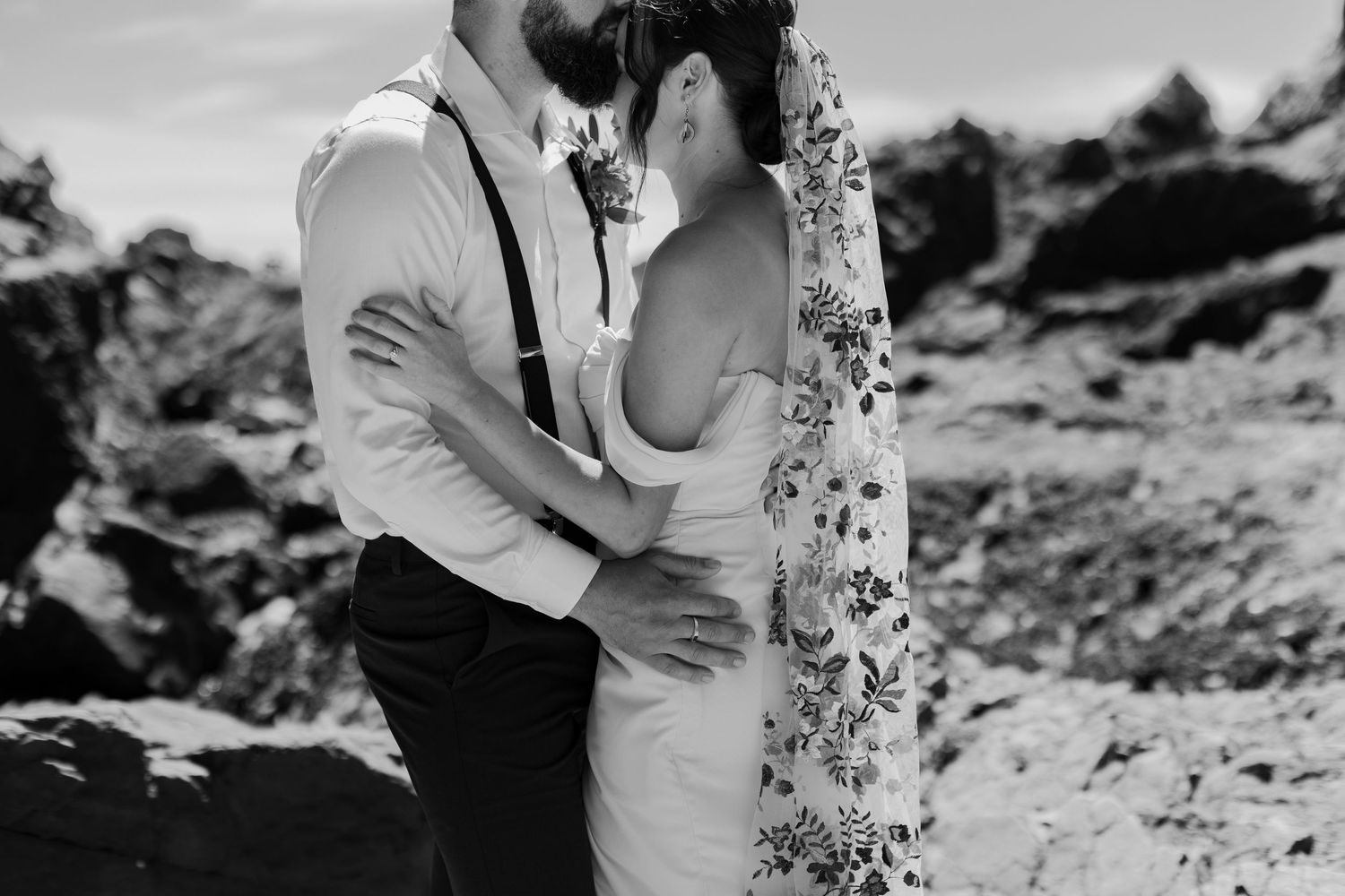 Black and white photo of bride and groom embracing intimately against rocky coastal scenery.