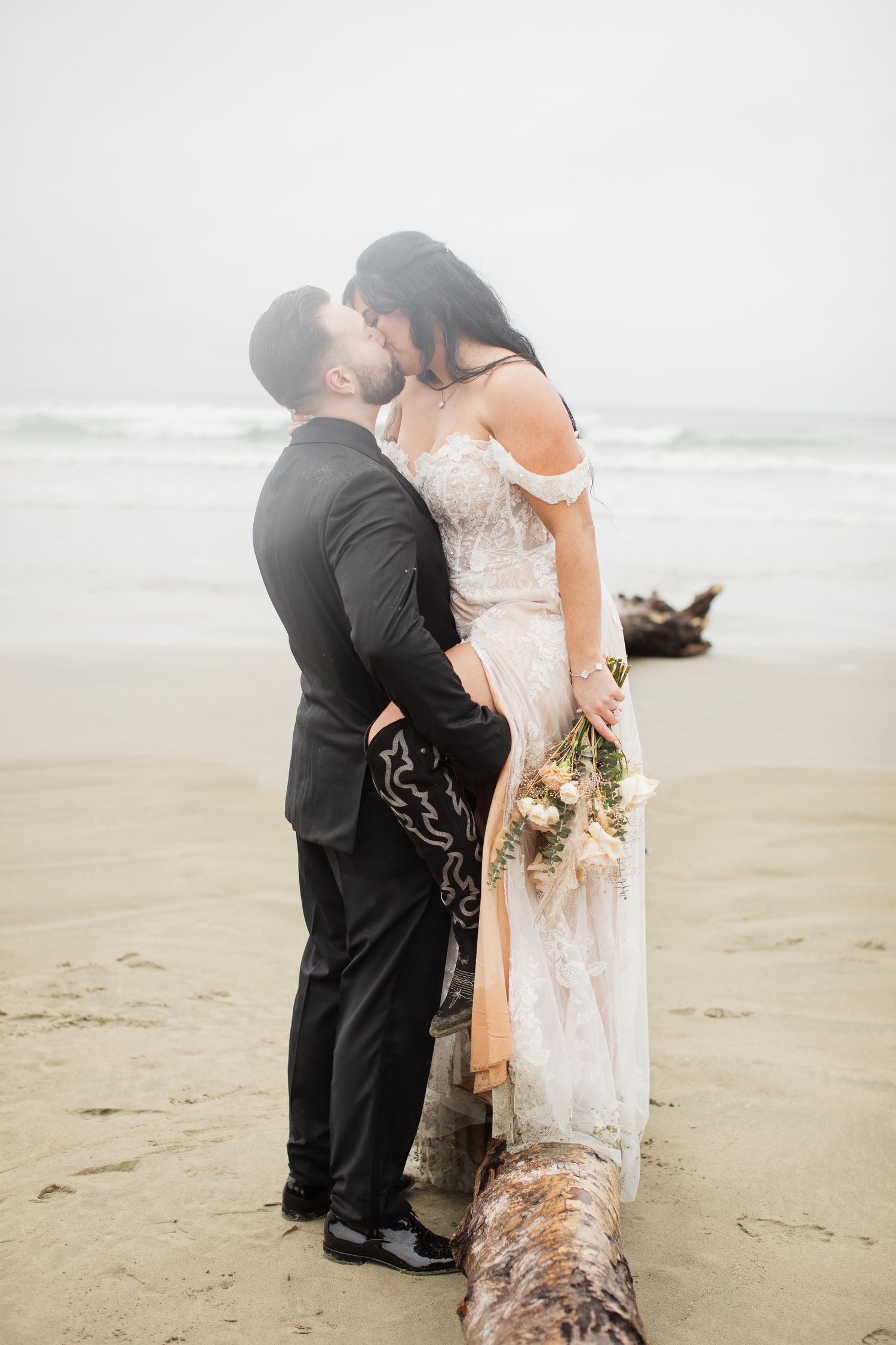 Bride and groom share romantic kiss on misty beach, bride holding boho bouquet near driftwood.