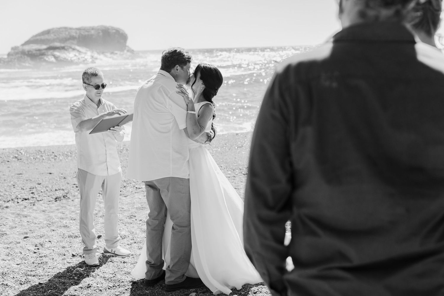Black and white photo of couple sharing first kiss at beach wedding ceremony with officiant and guests.