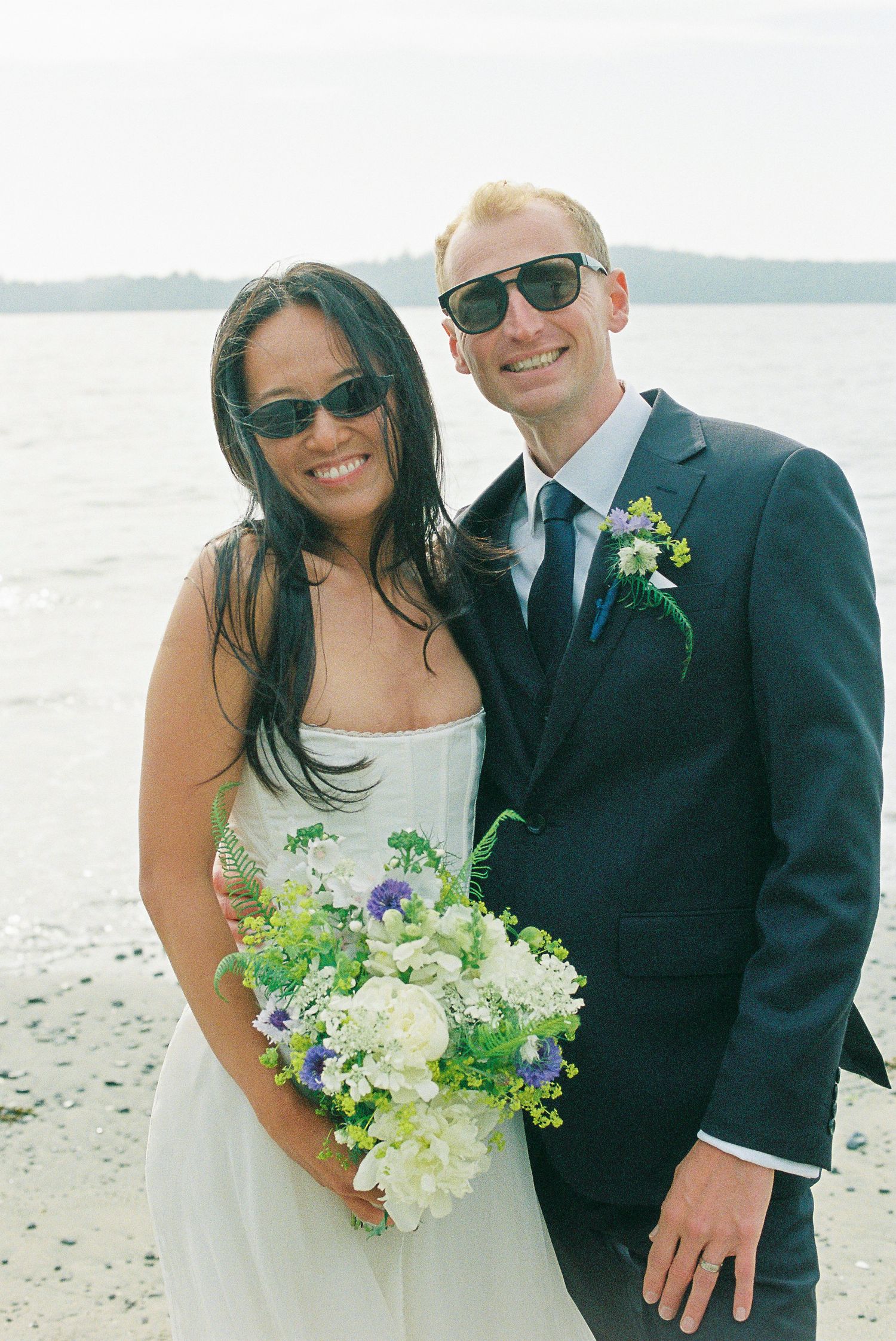 Smiling bride and groom wearing sunglasses posing on beach, bride holding purple and white bouquet.