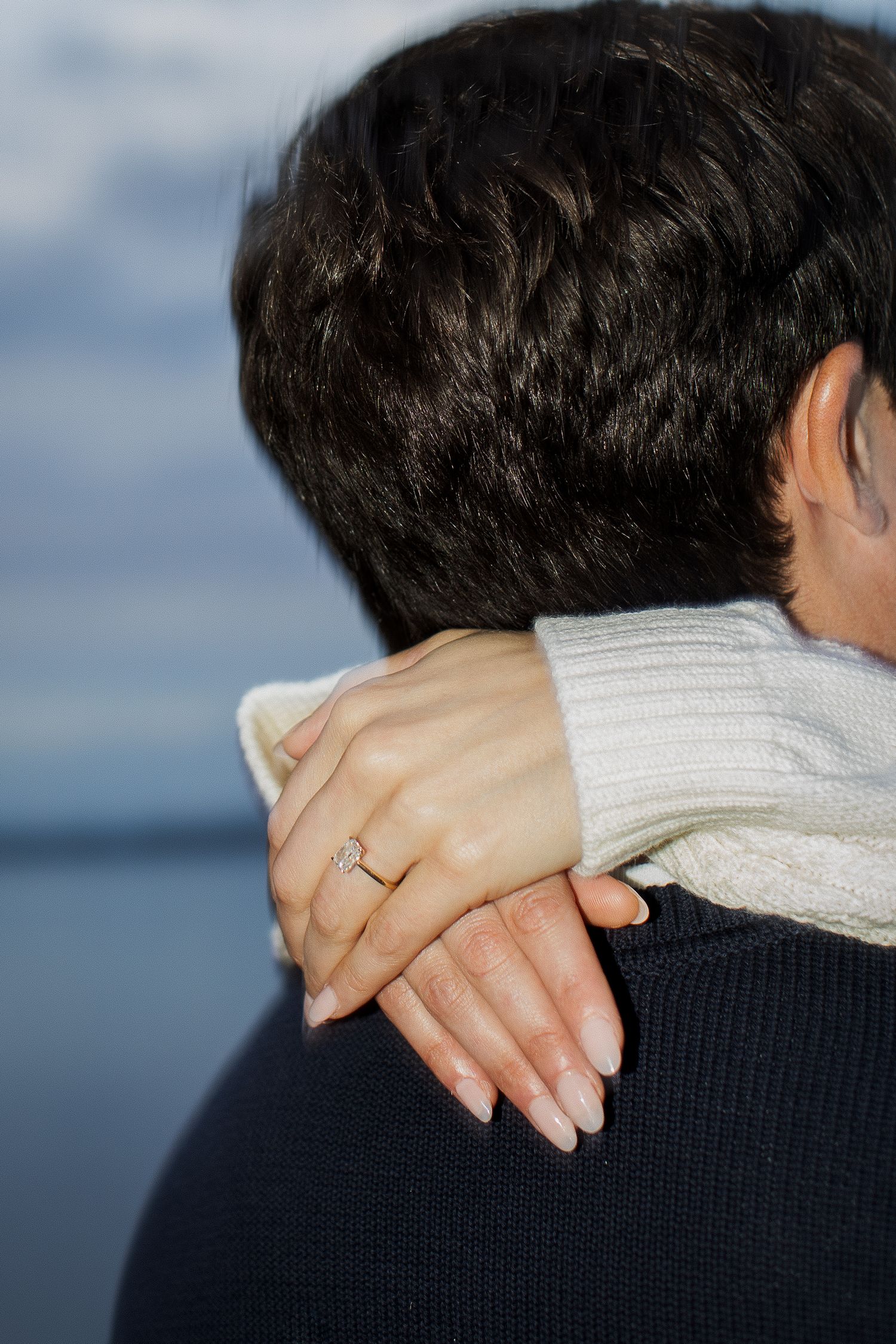 Close-up of a woman's hand with an engagement ring resting on a man's shoulder near the water.