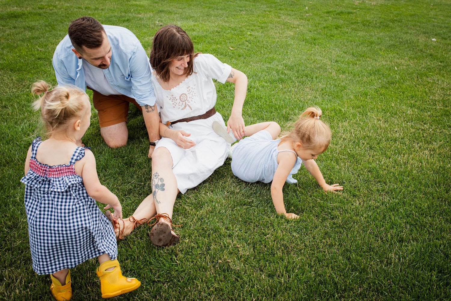 Family of four relaxing on green grass, parents sitting while two toddler girls play around them.