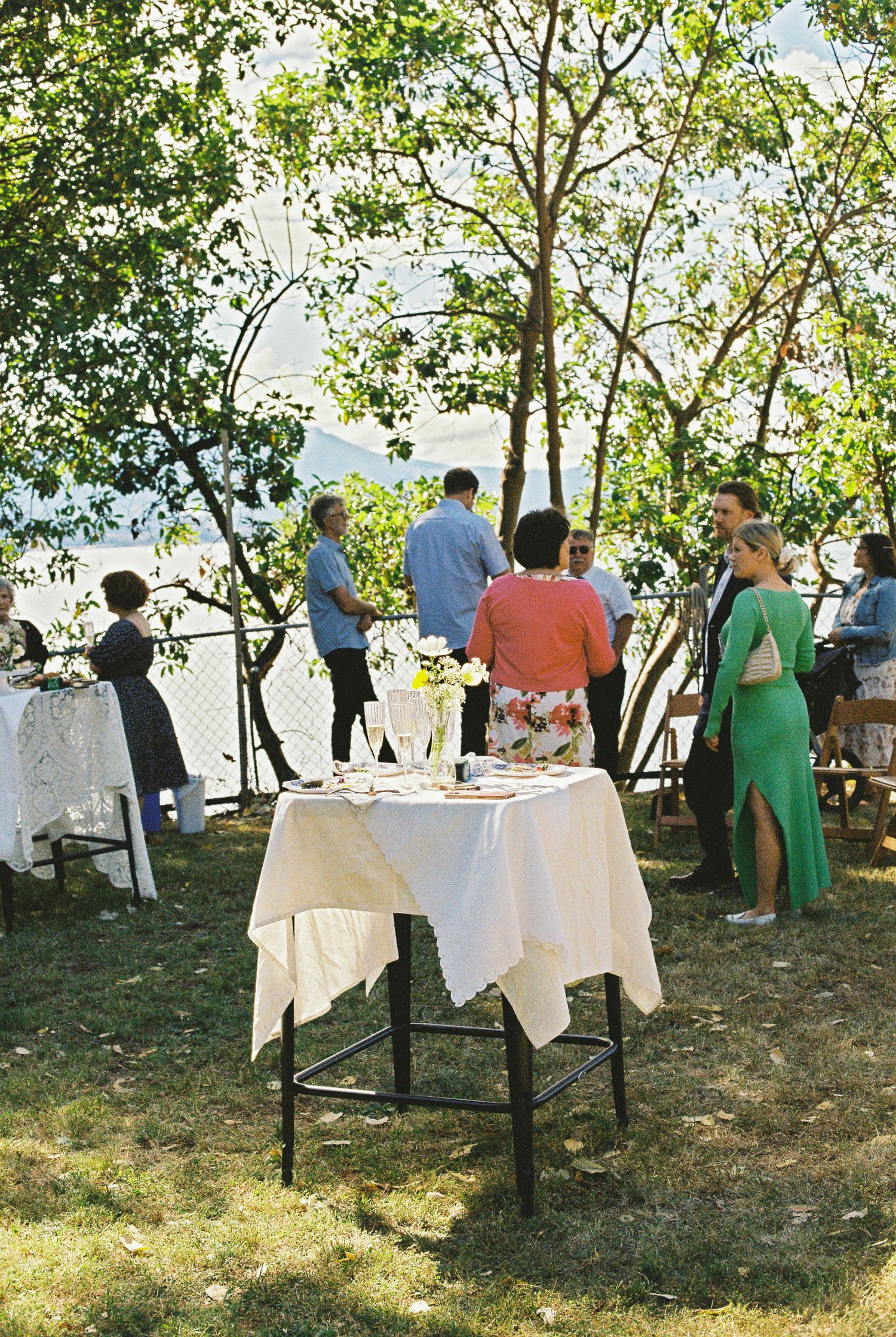 Outdoor wedding reception with guests mingling around white linen tables under green trees.