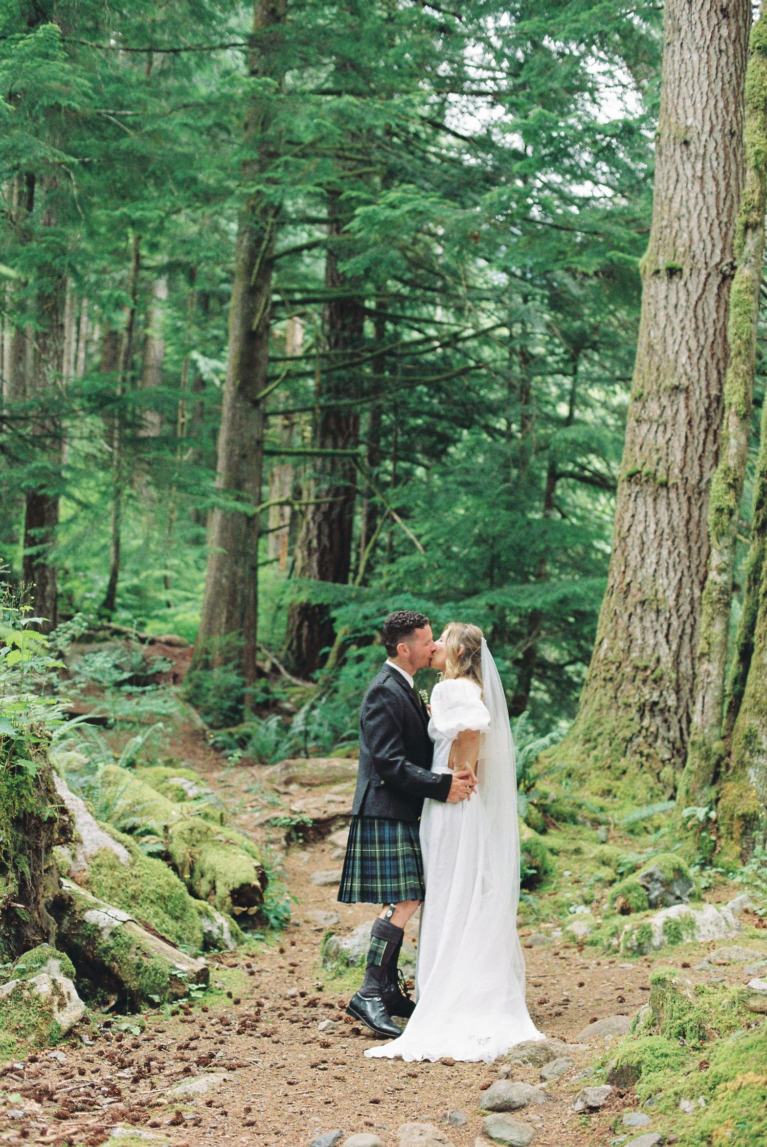 Bride in white gown and groom in tartan kilt share romantic kiss on mossy forest woodland path.