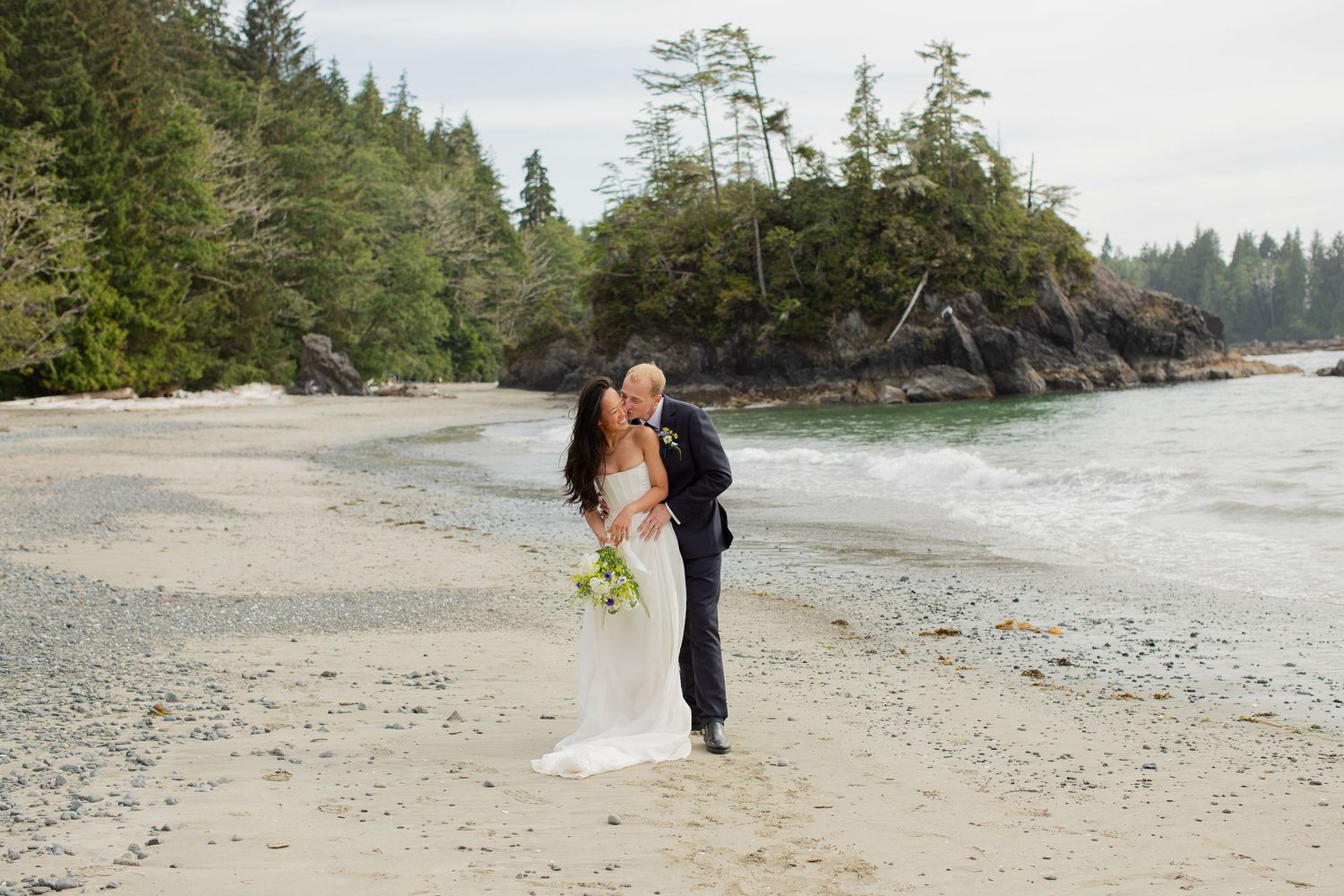 Bride and groom share a dip kiss on sandy beach with ocean backdrop.