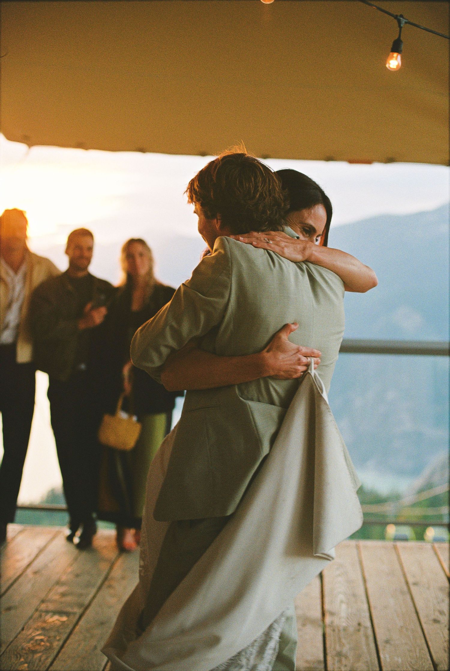 Newlyweds share first dance on mountain deck at sunset with guests watching and string lights above.