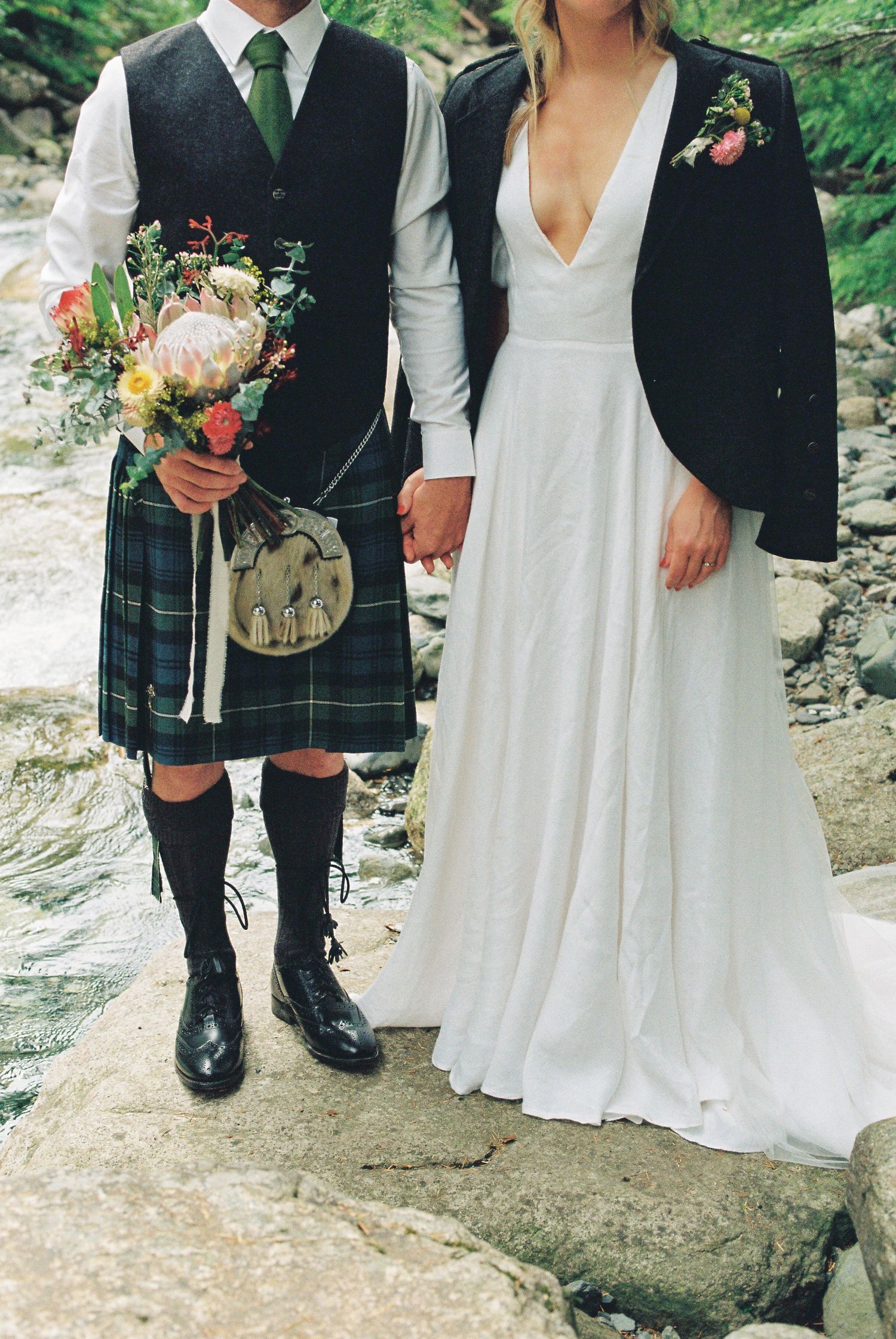 Scottish groom in tartan kilt holds bridal bouquet while holding hands with bride in white wedding gown.