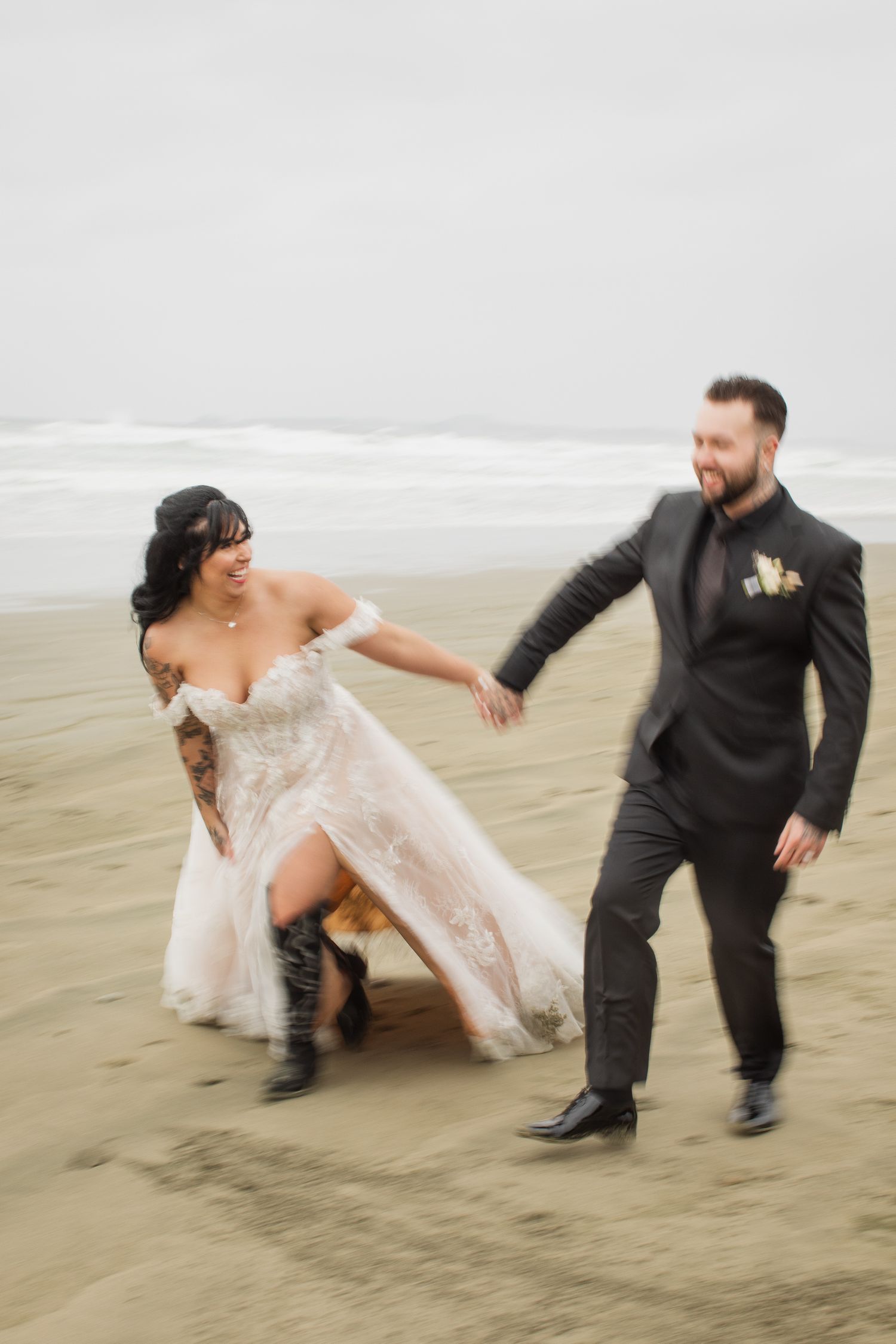 Bride in lace gown and cowboy boots dances playfully with laughing groom on sandy beach.