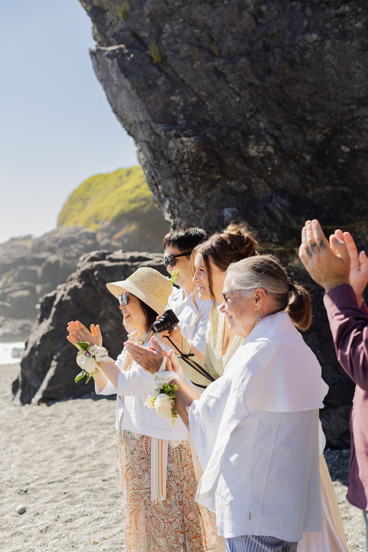 Wedding guests clap and celebrate after beach ceremony beside dramatic rock formations on a sunny day.