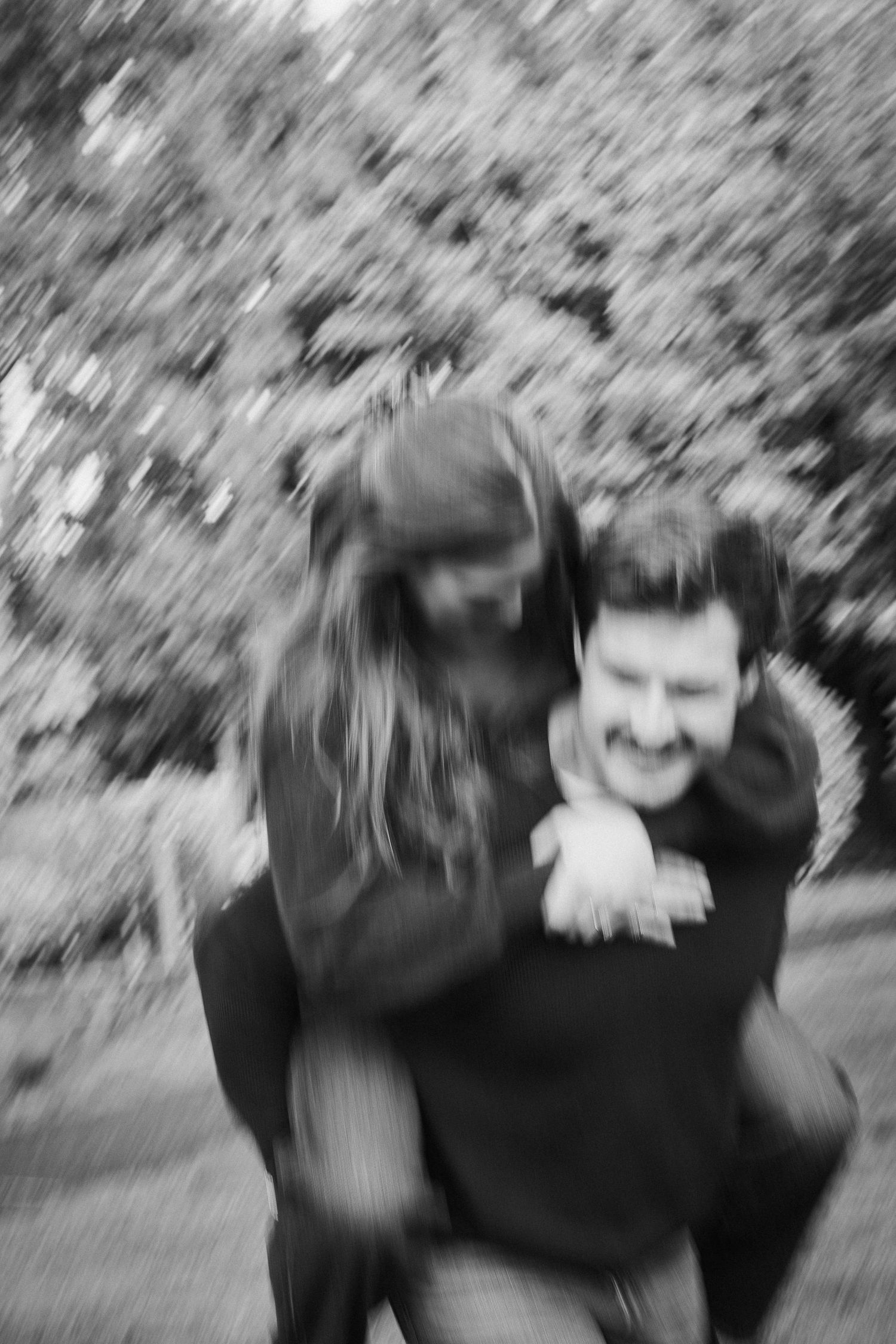 Blurred black and white photo of a laughing man giving a woman a piggyback ride outdoors.