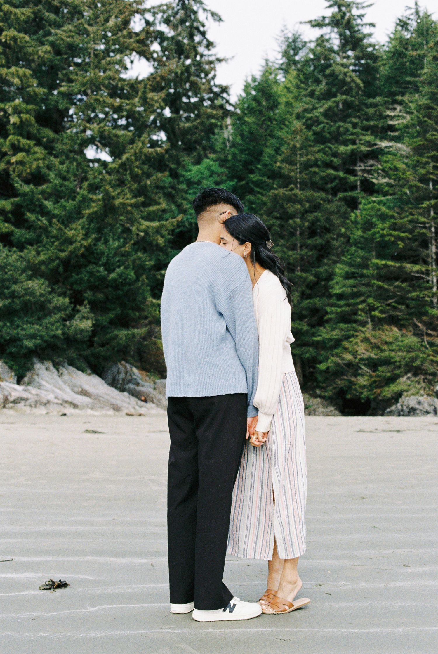 Romantic couple standing close together holding hands on a beach with evergreen trees.