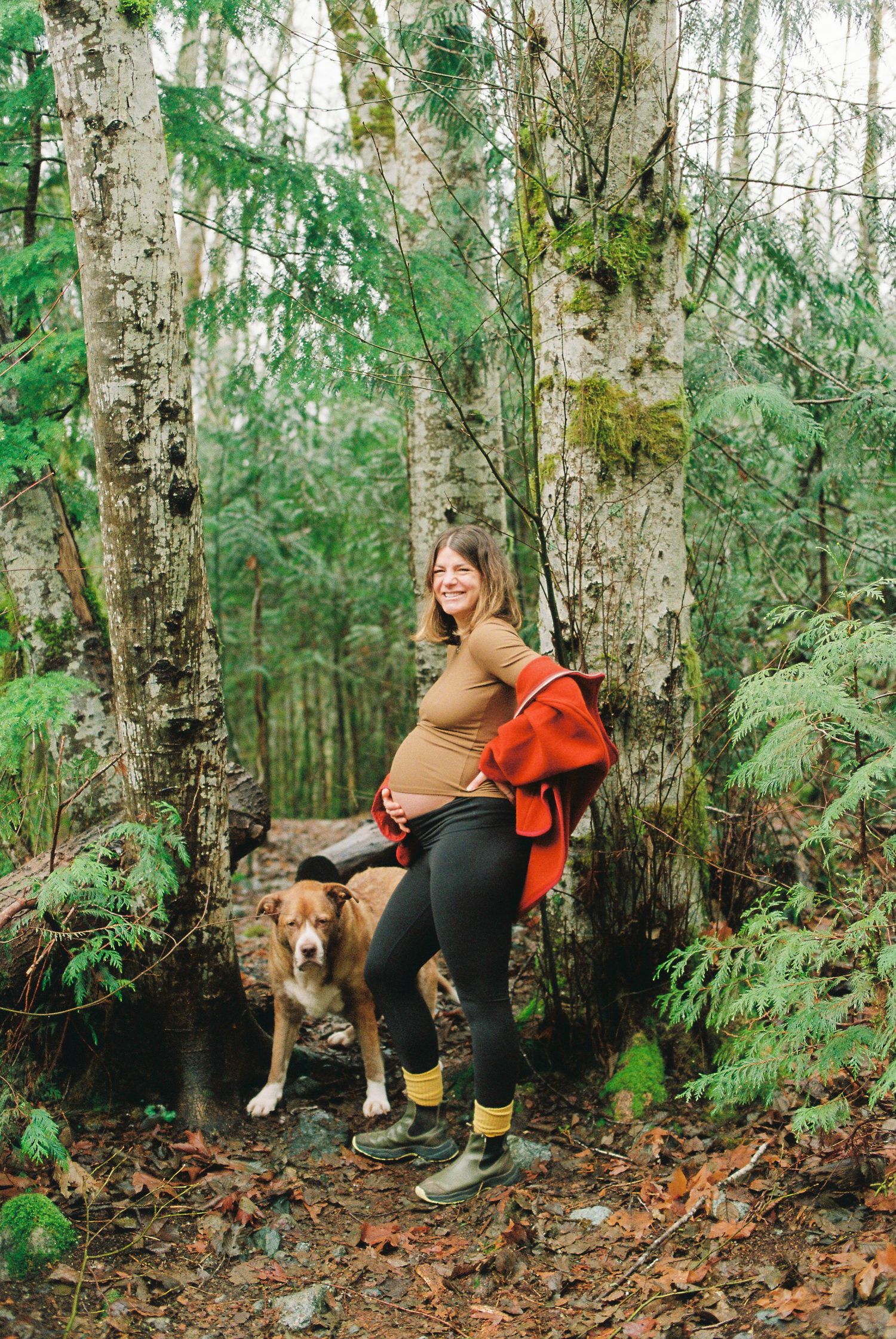 Happy pregnant woman posing with golden retriever dog in mossy Pacific Northwest forest.