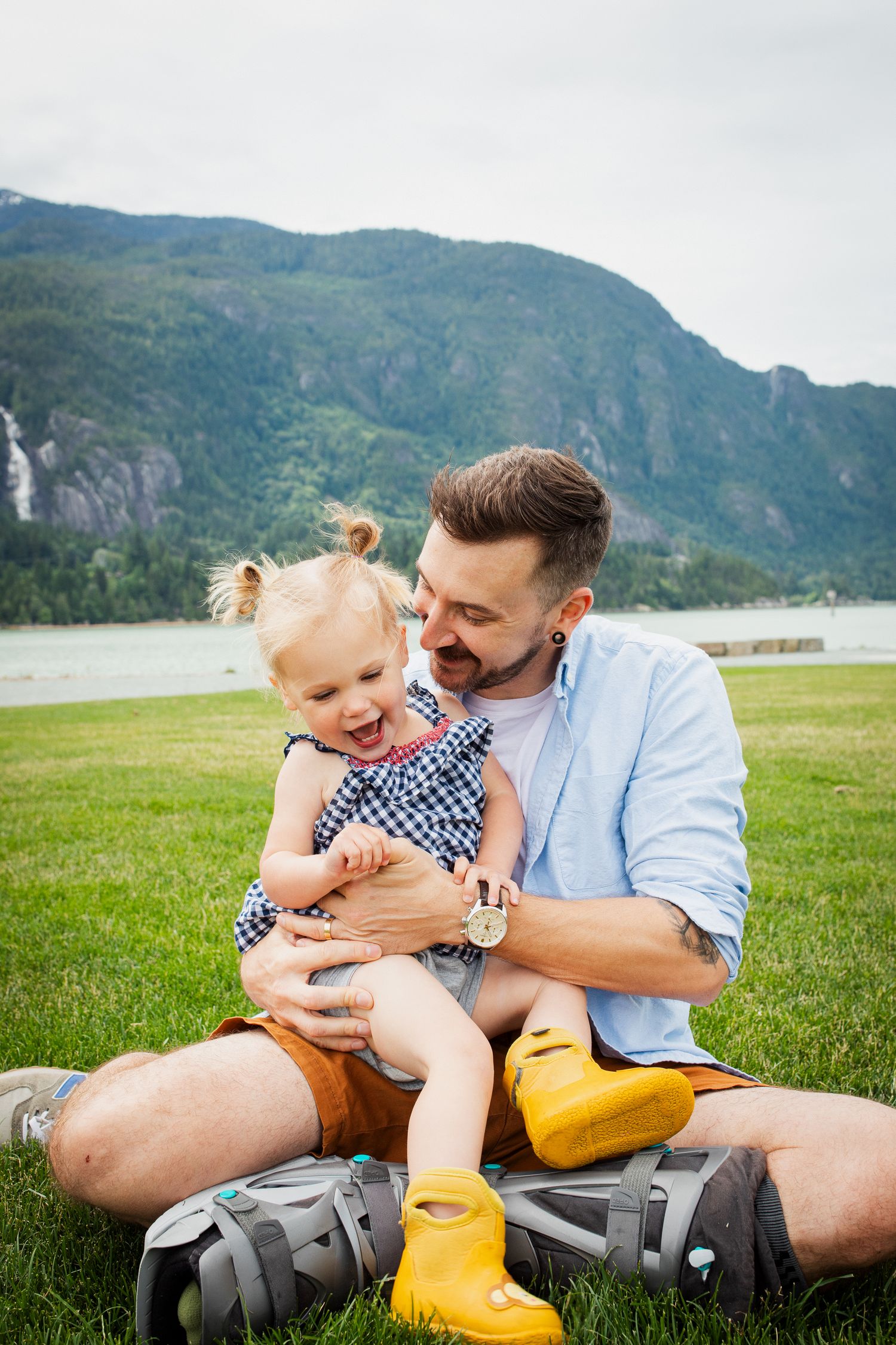Father sitting on grass holding laughing toddler daughter with mountain and lake scenery behind them.