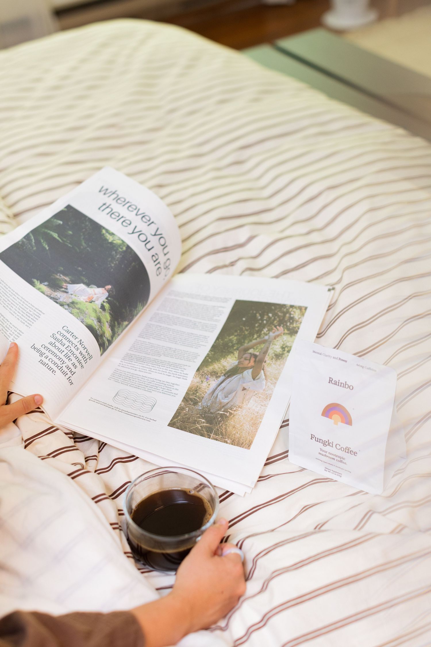 Person holding black coffee cup reading a magazine in bed with a Pangia Coffee packet nearby.