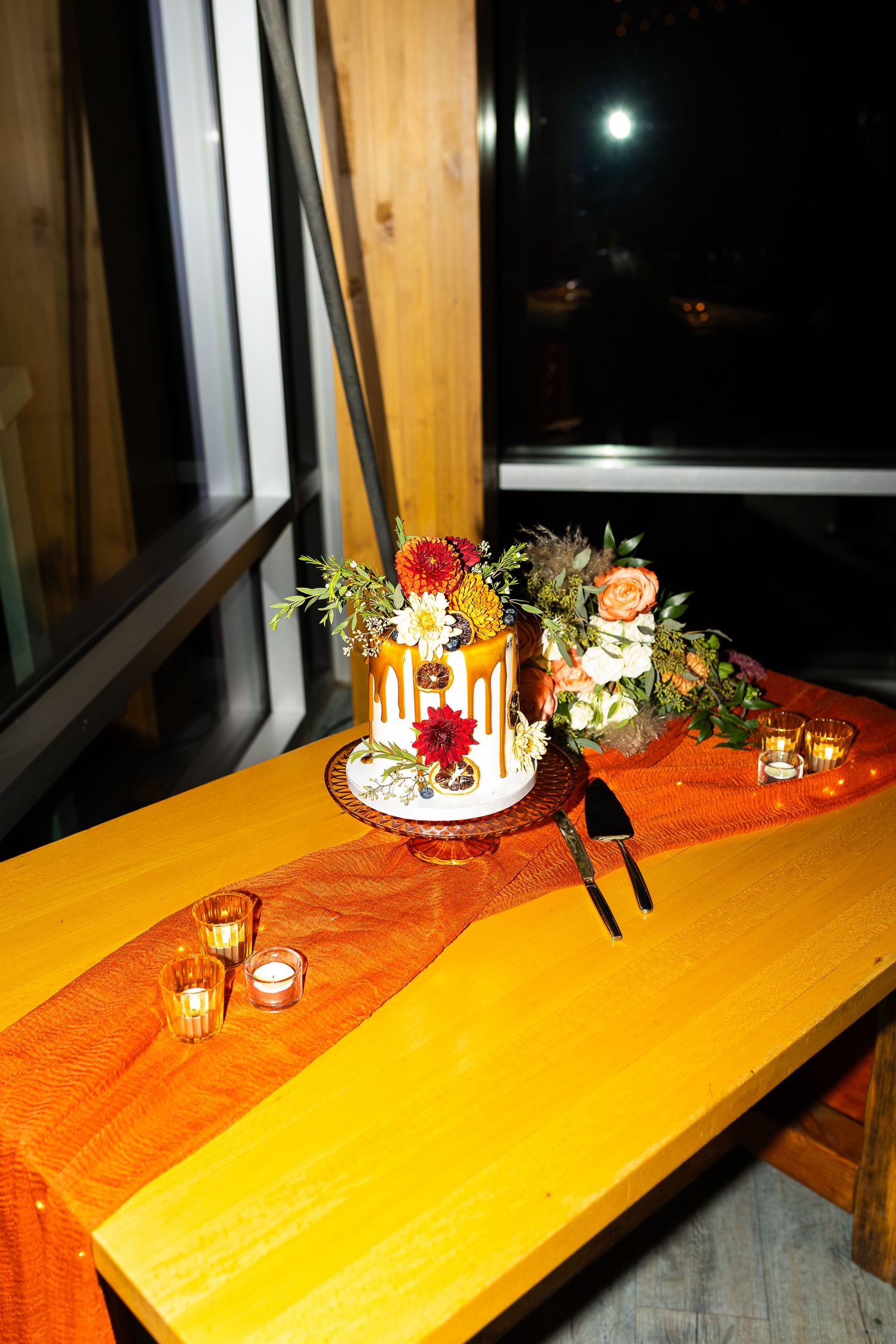 Elegant one-tier wedding cake with caramel drip and floral topper on orange table runner with candles at night.