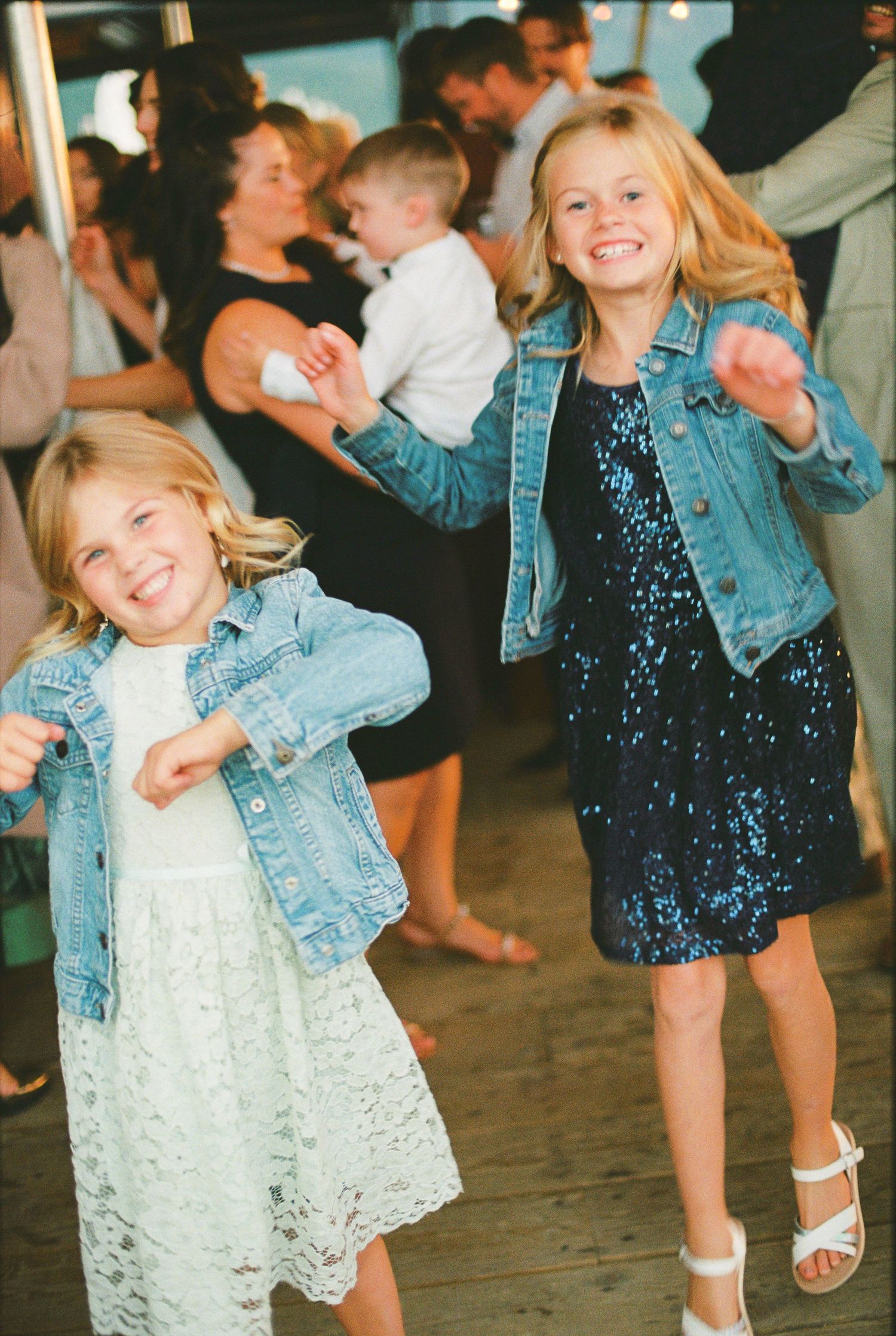 Two young flower girls in denim jackets dance joyfully at wedding reception on decorated dance floor.