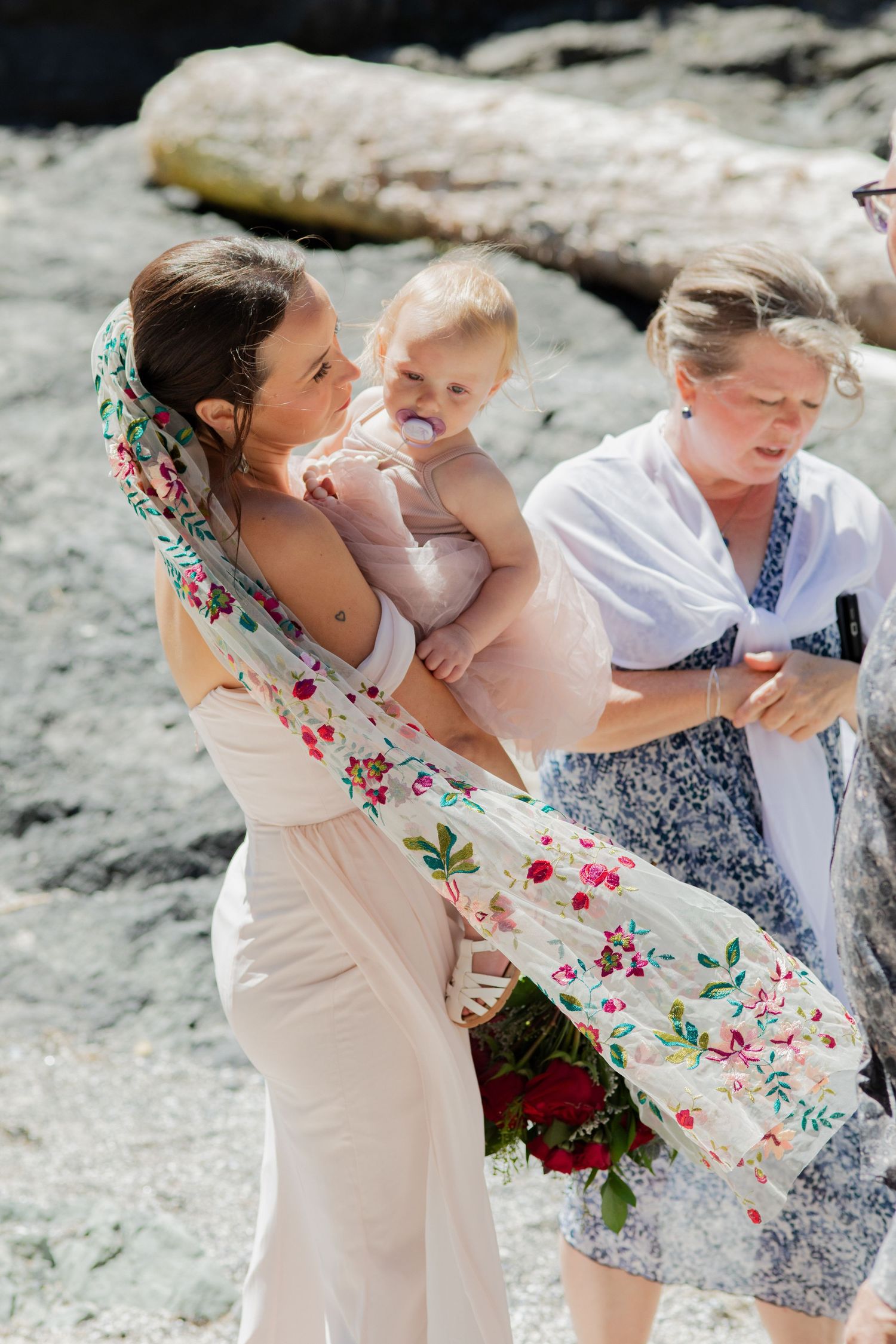 Bride in blush dress with floral shawl holds baby while older woman assists at beach wedding.