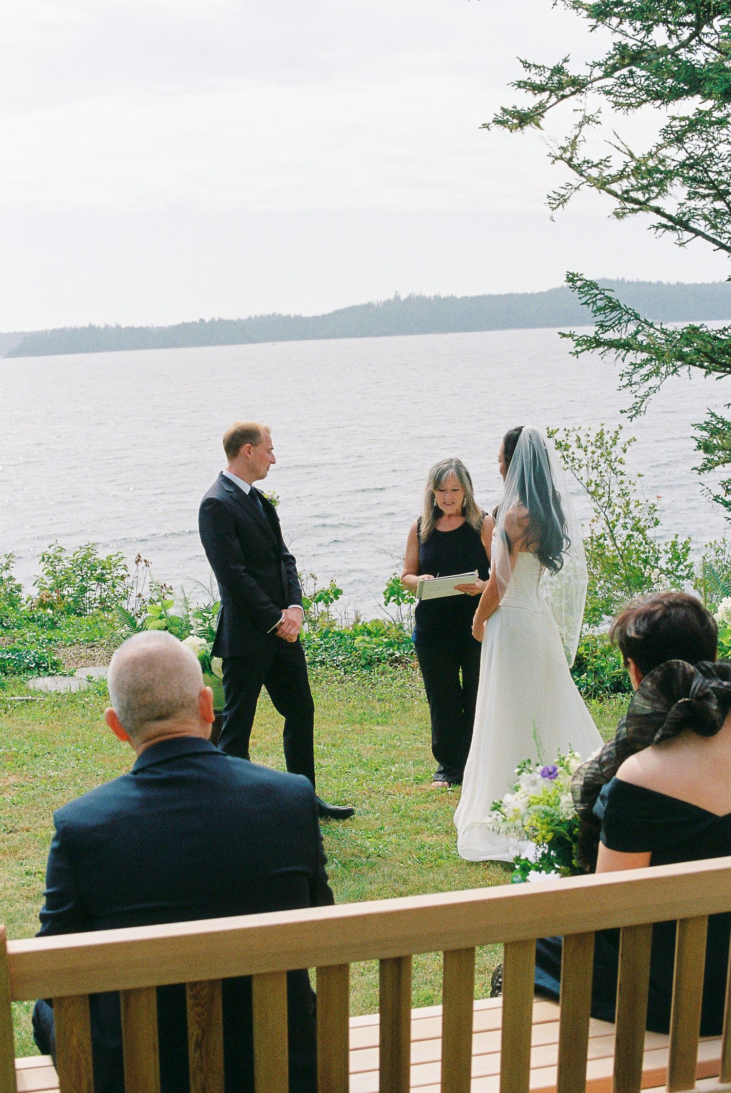 Intimate outdoor wedding ceremony overlooking calm ocean waters with officiant and small guest gathering.