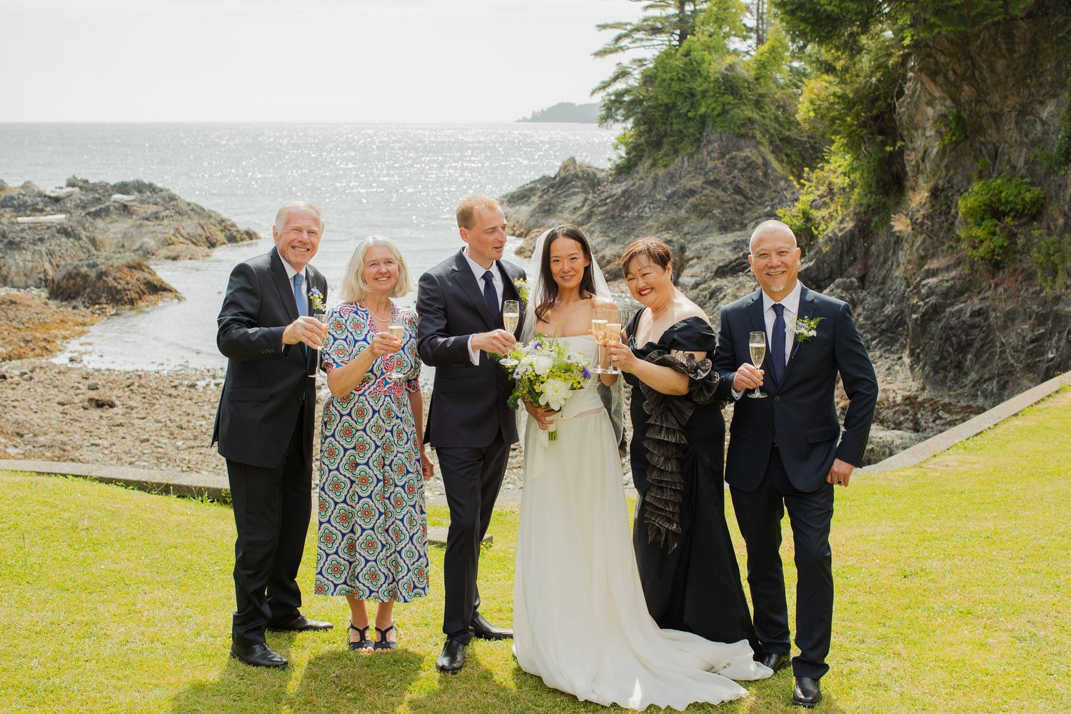 Wedding party group photo on green lawn with scenic ocean and rocky coastline backdrop.