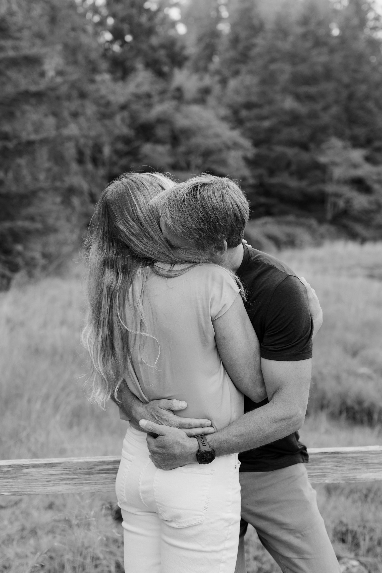 Black and white photo of a couple sharing an intimate embrace near a wooden fence in a natural setting.