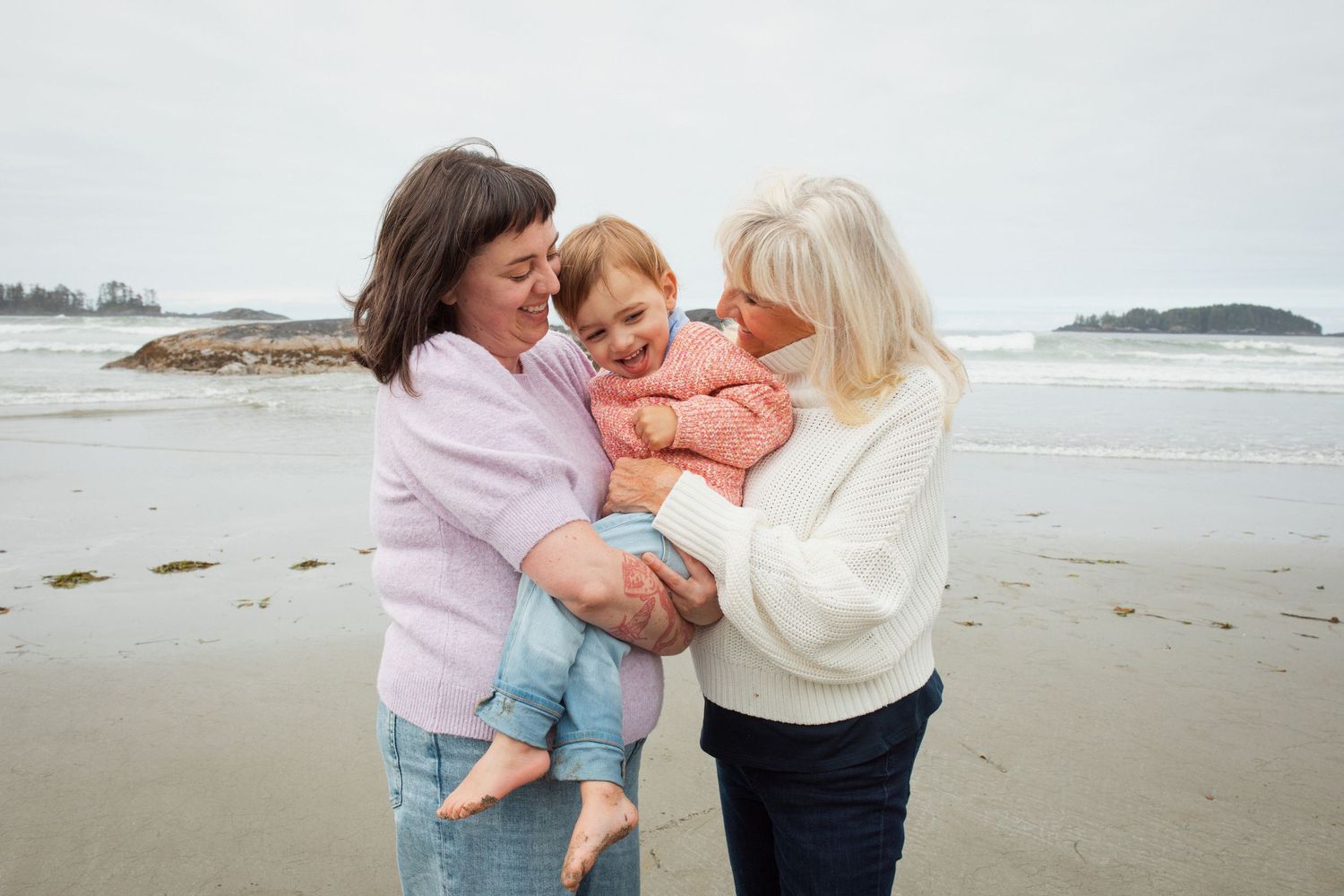 Two women share a joyful moment holding a smiling toddler on a cloudy beach shoreline.