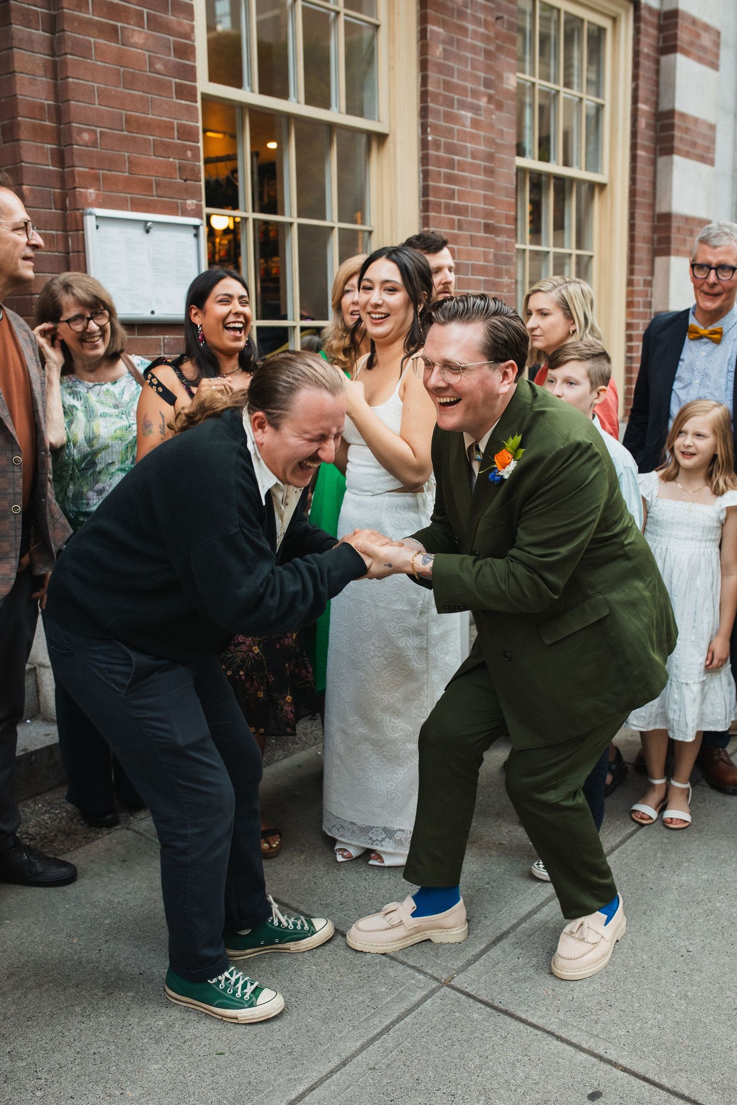 Two men dancing and celebrating joyfully outside a brick building at a lively wedding reception.