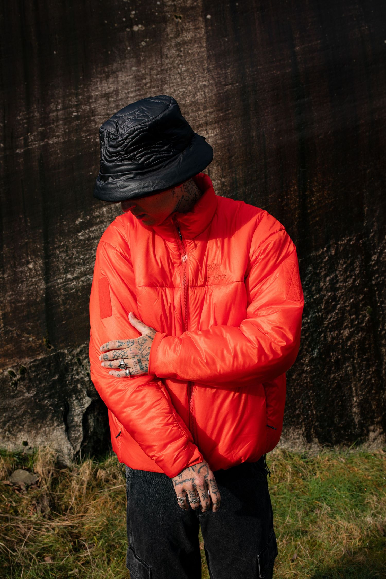 Tattooed man in red puffer jacket and black bucket hat standing against dark stone wall.