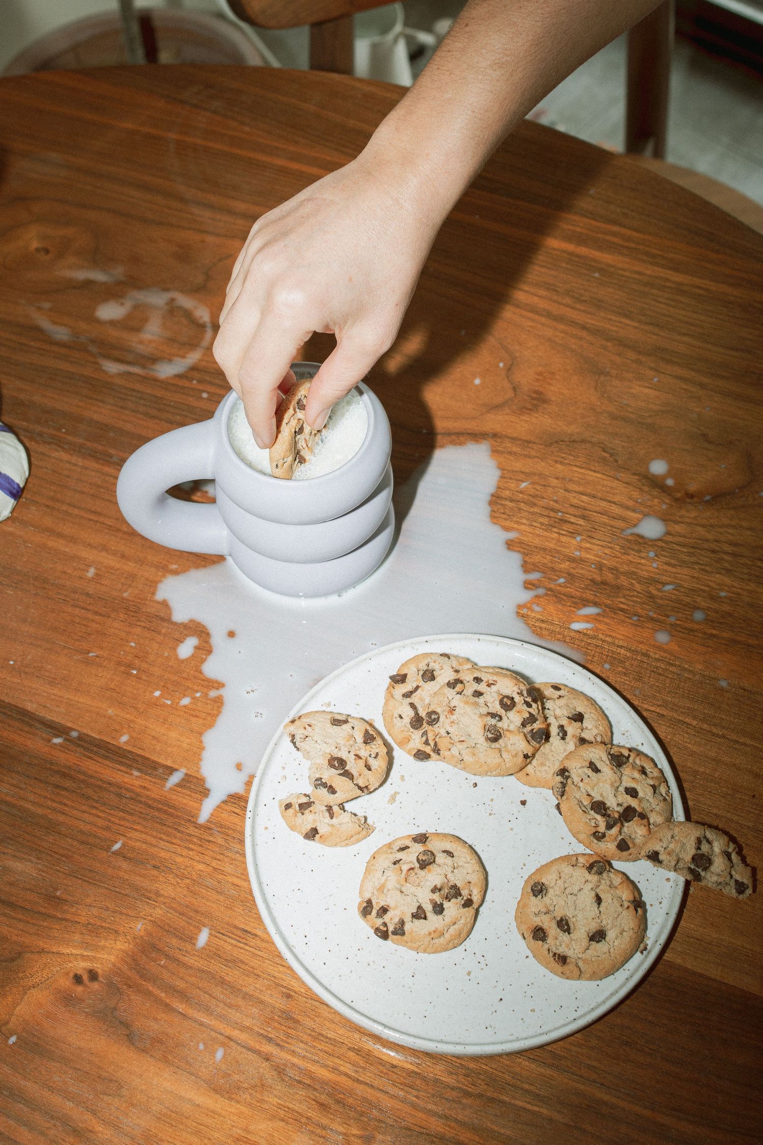 Hand dunking a chocolate chip cookie into a stacked ceramic mug with spilled milk on wood table.