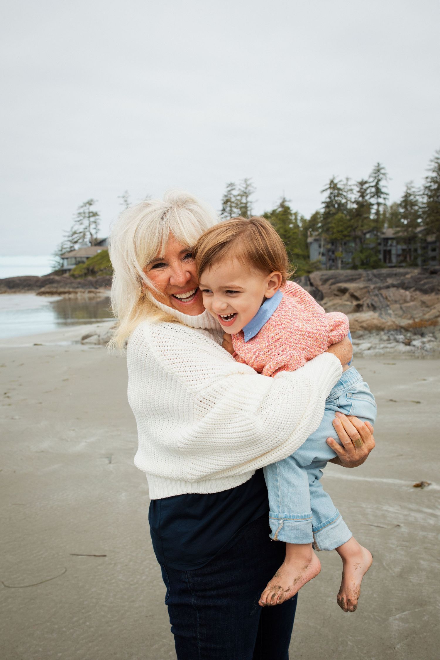 A smiling grandmother holds a laughing toddler on a sandy beach with rocky coastline in background.