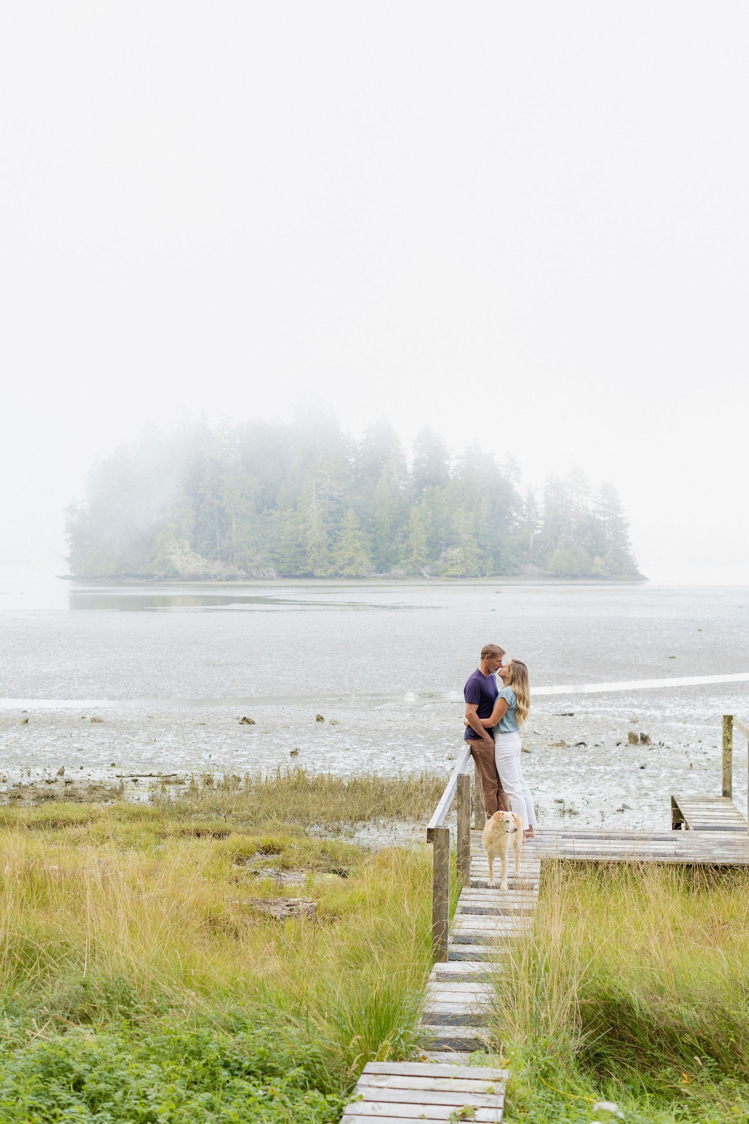 A couple shares a romantic embrace on a wooden dock overlooking a foggy island landscape with their dog.