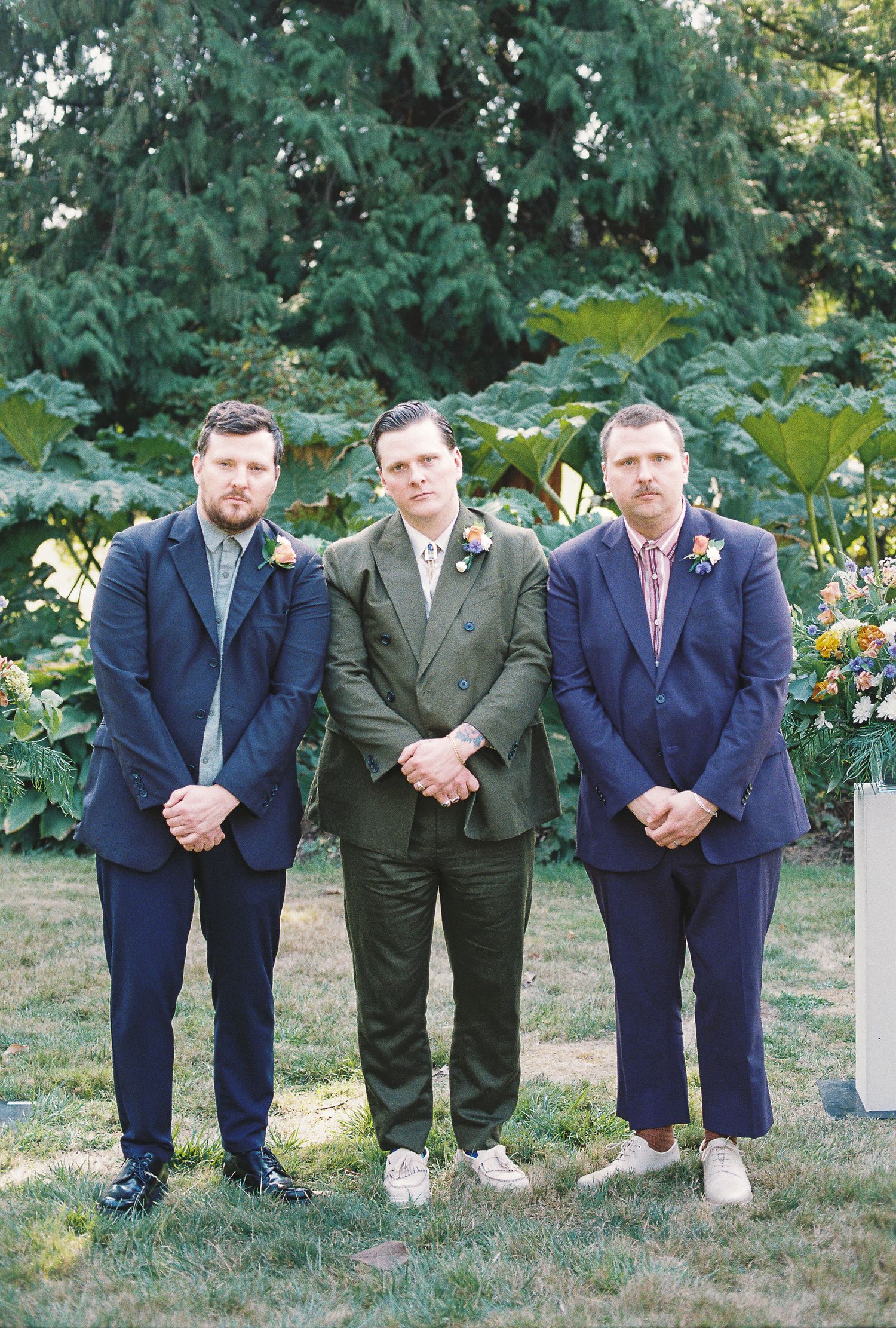 Groom in olive suit stands between two groomsmen in navy suits at garden wedding.
