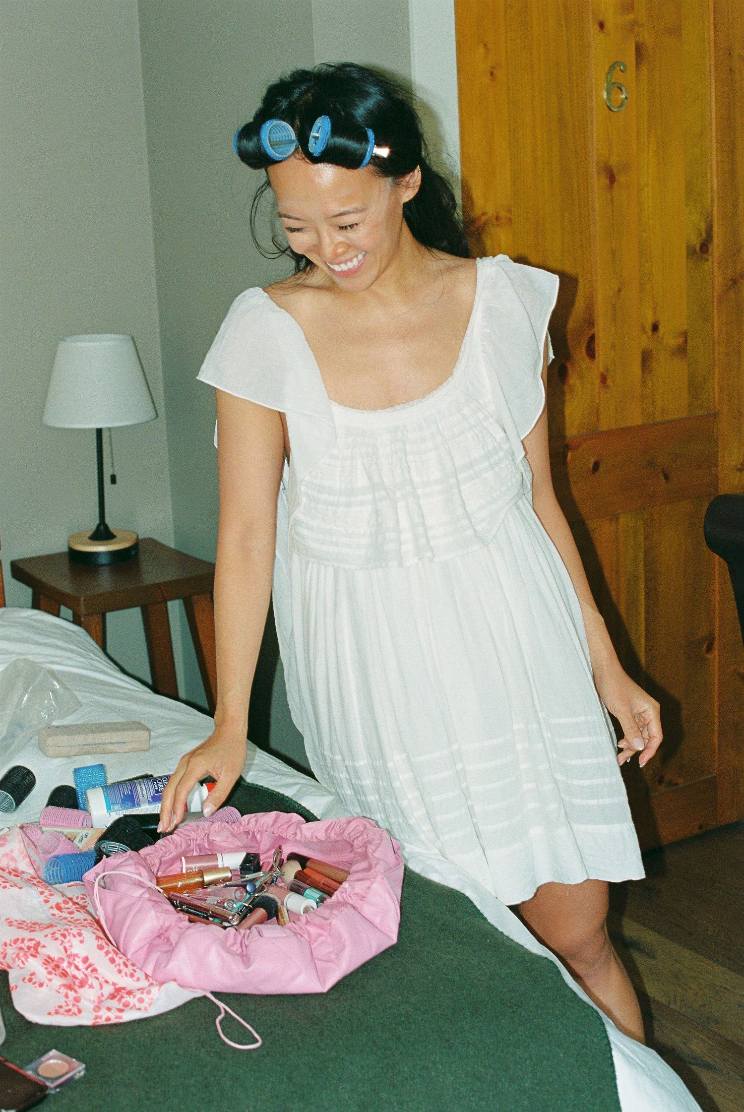 Bride smiling in white dress with blue hair rollers, looking at pink makeup bag on bed.