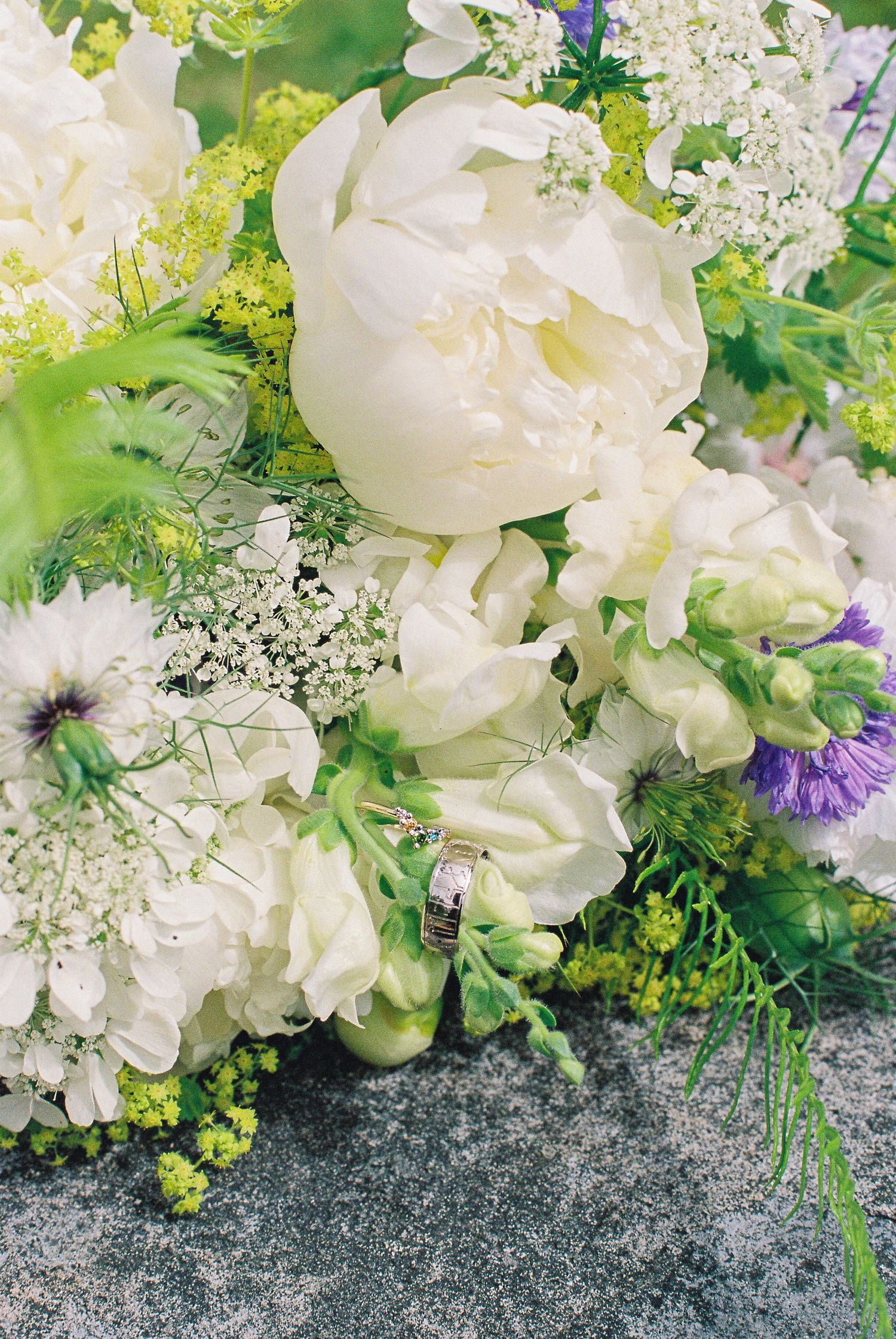 Lush bridal bouquet featuring white peonies, anemones, sweet peas, and greenery with wedding rings.