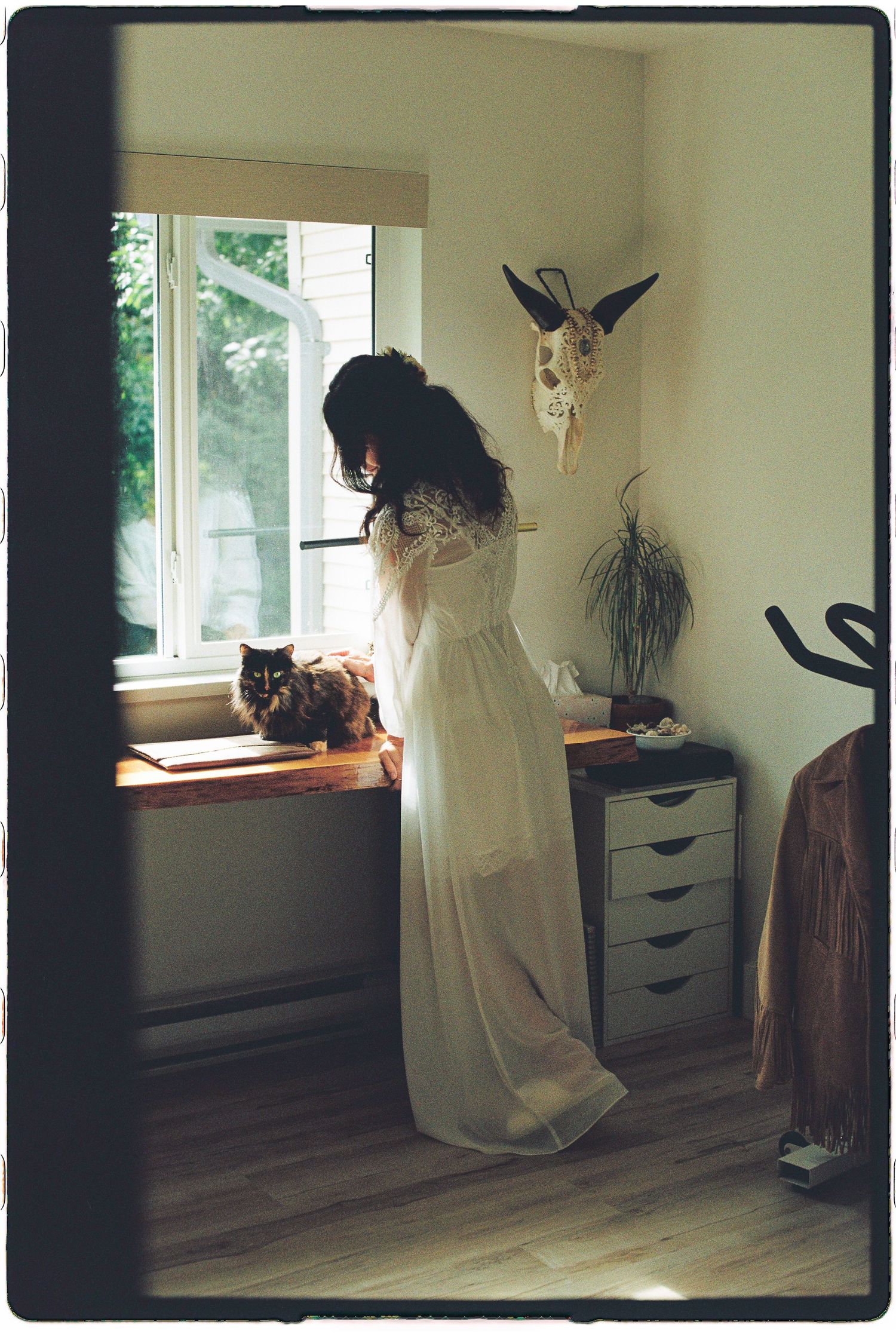 Bride in flowing white gown stands by window with cat on dresser beside animal skull wall decor.