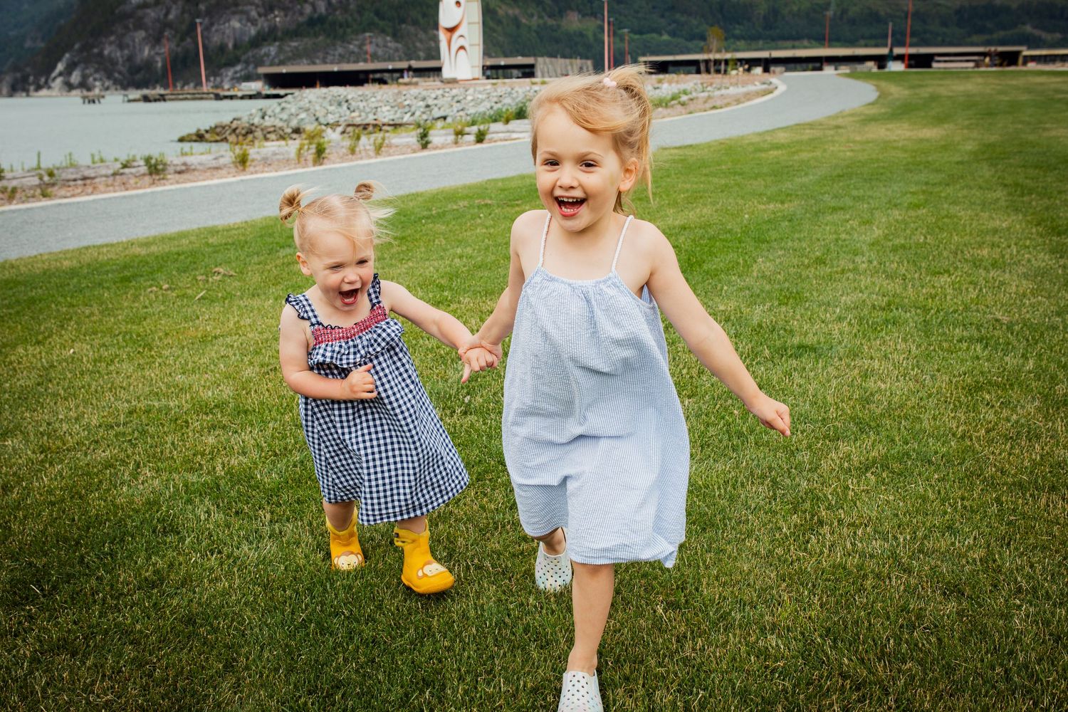 Two young girls in dresses holding hands and running on green grass near a lake.