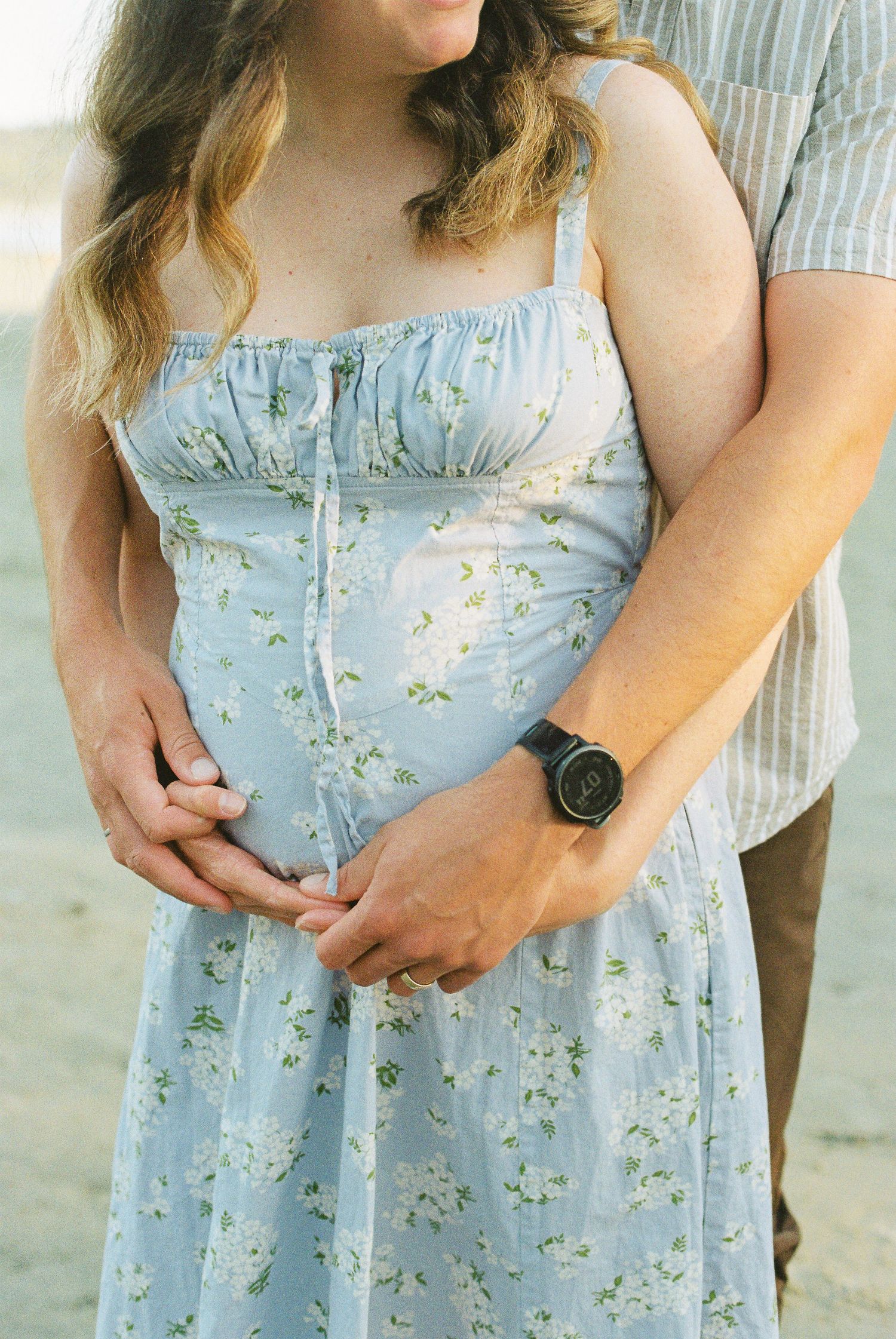 Close-up of couple's hands gently holding pregnant belly in a blue floral maternity dress on the beach.