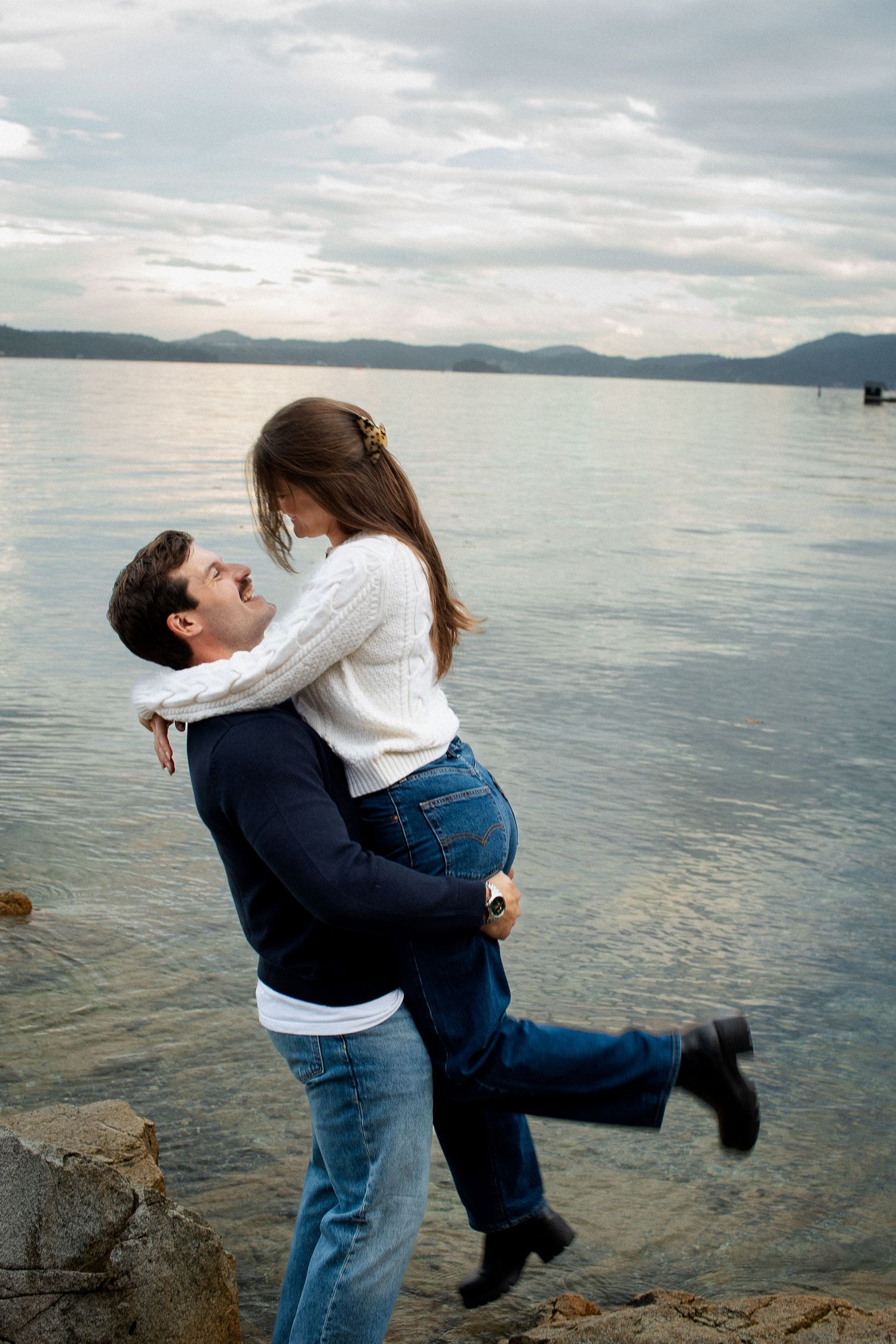 Man lifting woman in joyful embrace by rocky lakeside under dramatic cloudy sky.