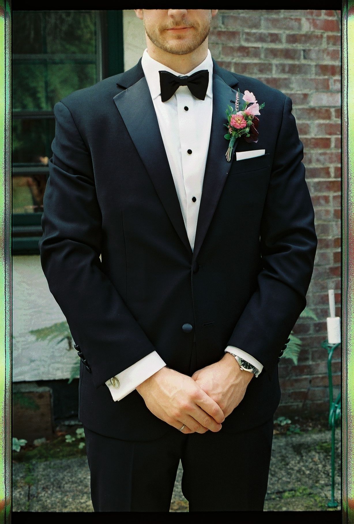 Groom in black tuxedo with bow tie and pink boutonniere stands clasping hands before a brick wall.