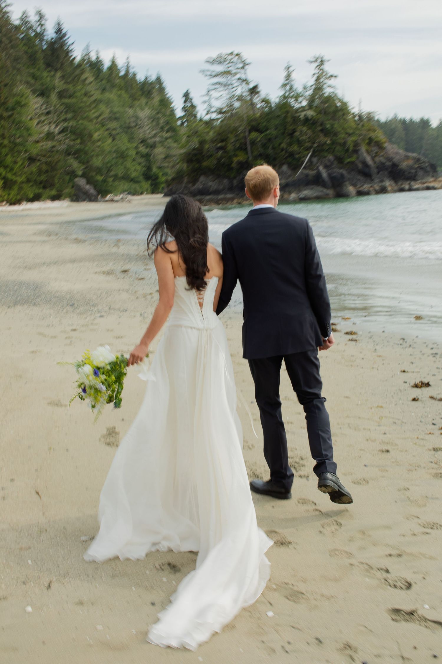 Bride holding bouquet and groom walk hand in hand along a sandy beach with forested shoreline behind.