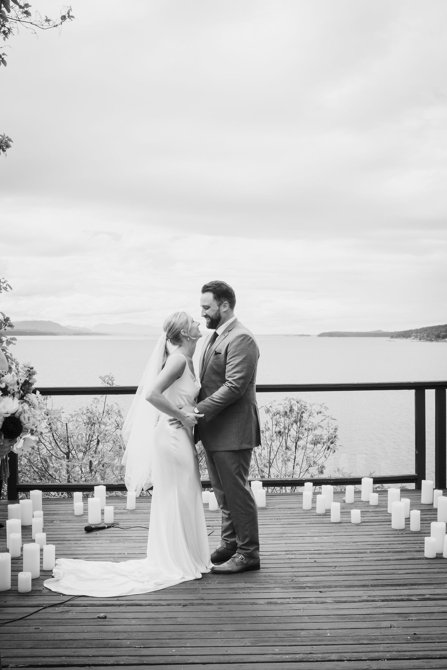 Black and white photo of bride and groom exchanging vows on a candle-lit lakeside deck.