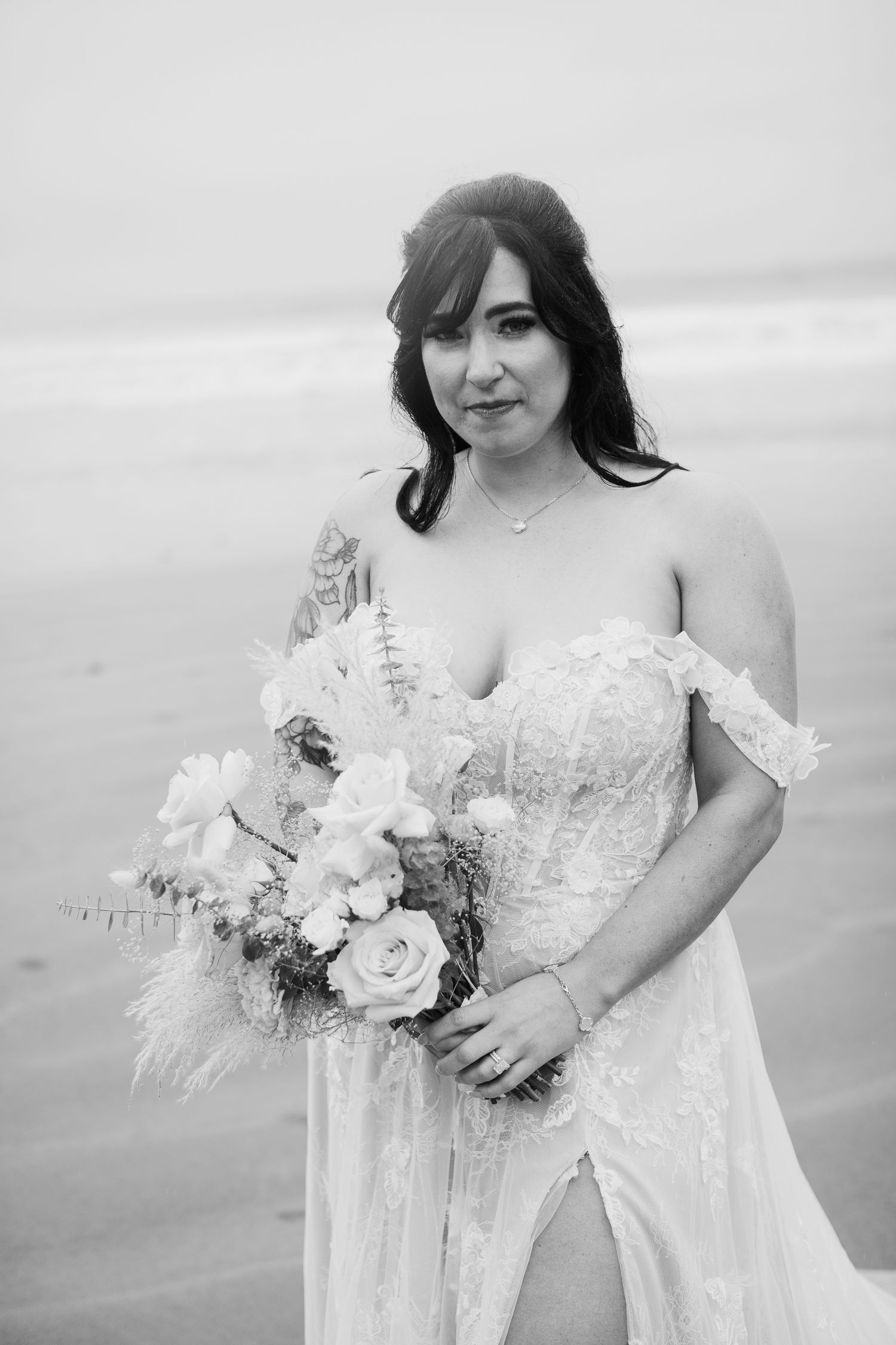 Bride in lace off-shoulder gown holds boho bouquet on misty beach in black and white portrait.