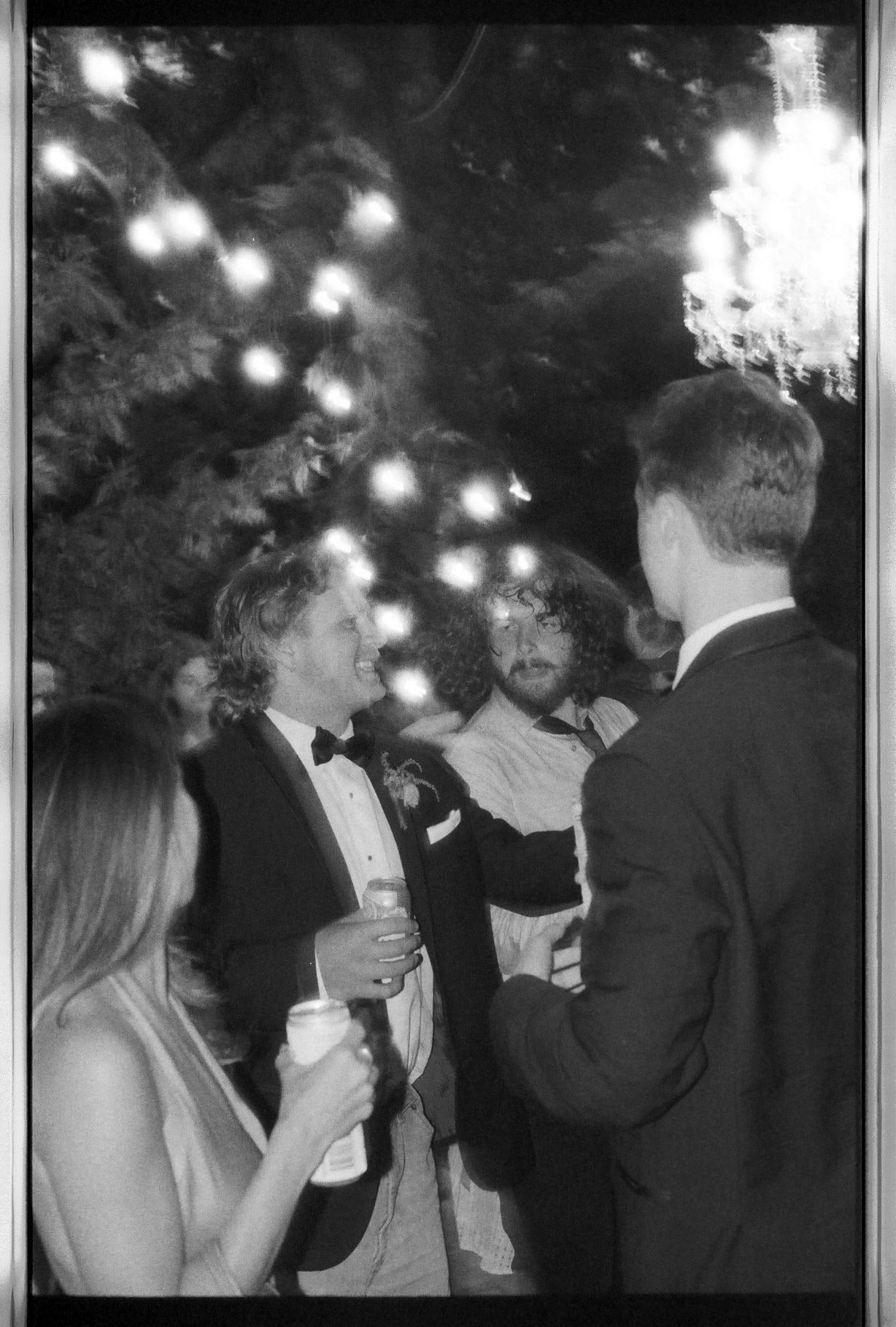 Black and white photo of wedding guests socializing under string lights at an outdoor evening reception.