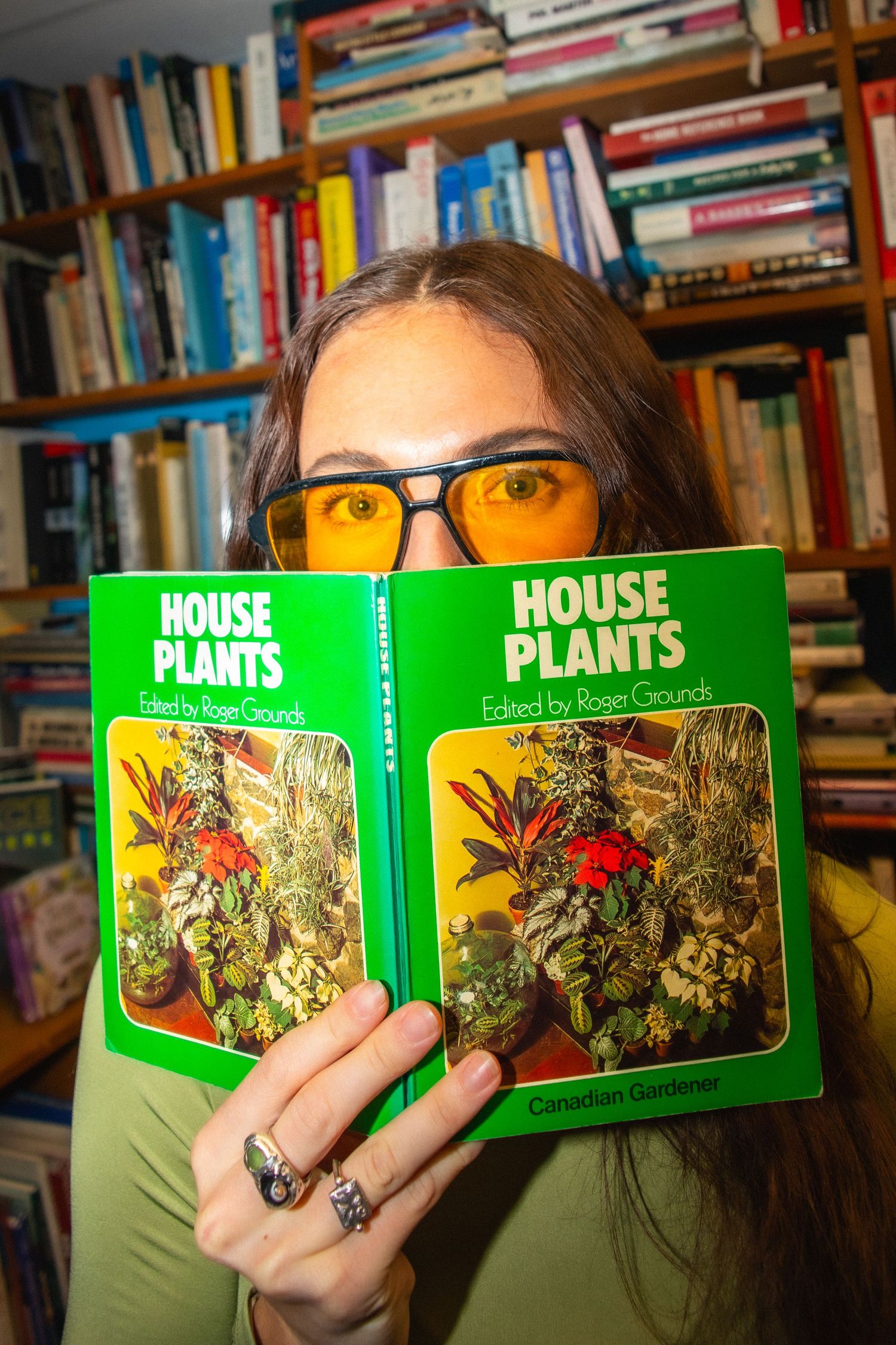 Woman holding a House Plants book edited by Roger Grounds in front of her face, wearing yellow tinted glasses.