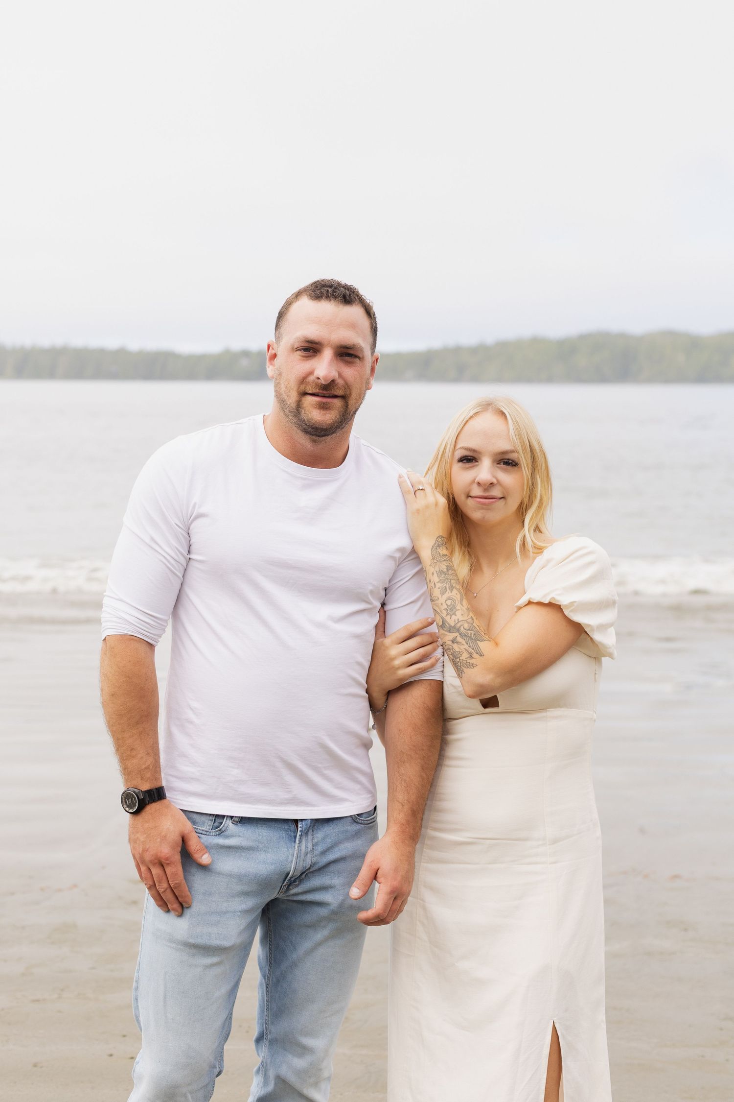 Couple posing together on a beach, woman in white dress holding man's arm, trees in background.